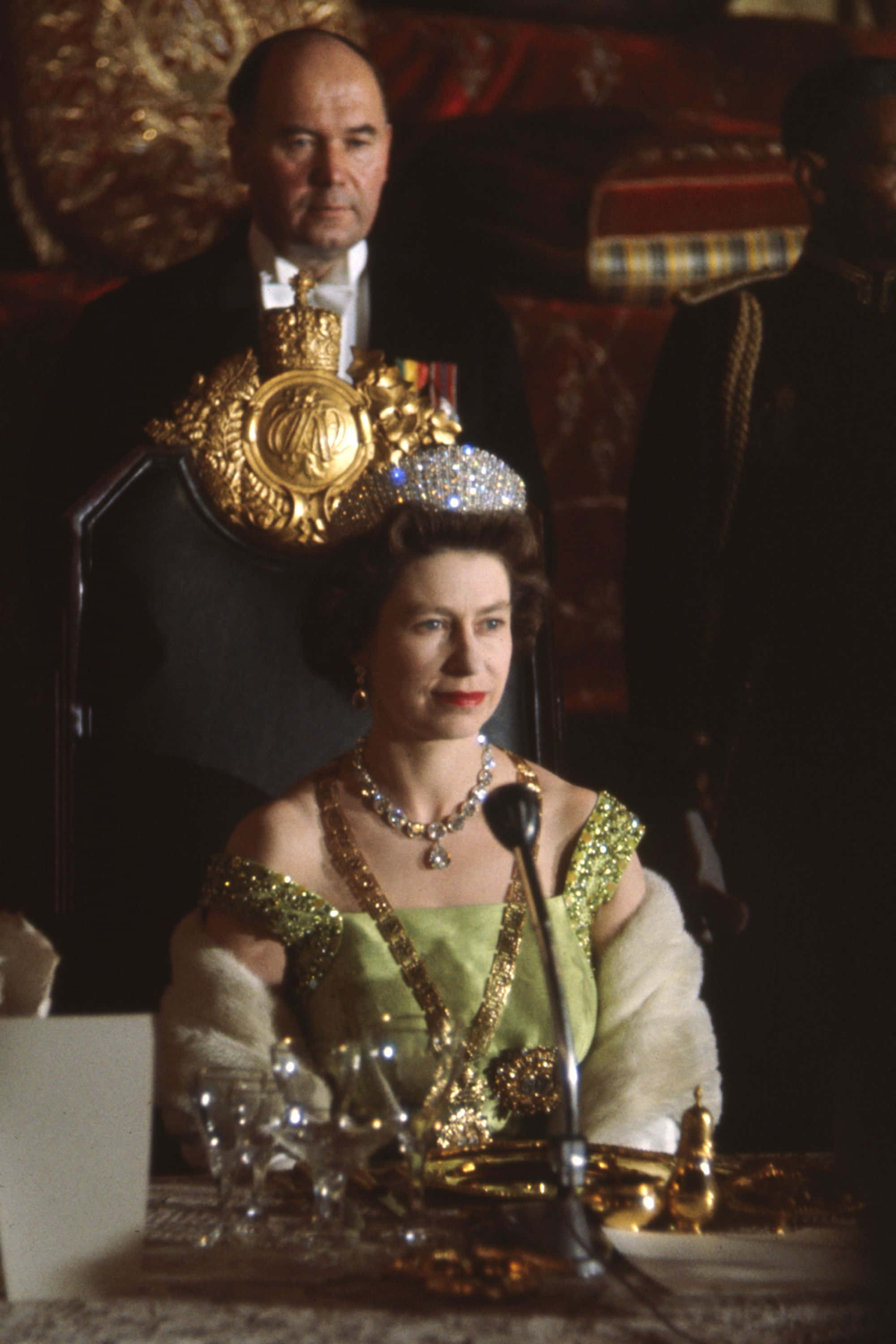 Queen Elizabeth 桌布 壁紙 — 伊利沙伯二世女王身穿綠色長袍和鑽石頭飾，端莊地坐在正式活動中，身後椅子上放著一頂金色王冠。