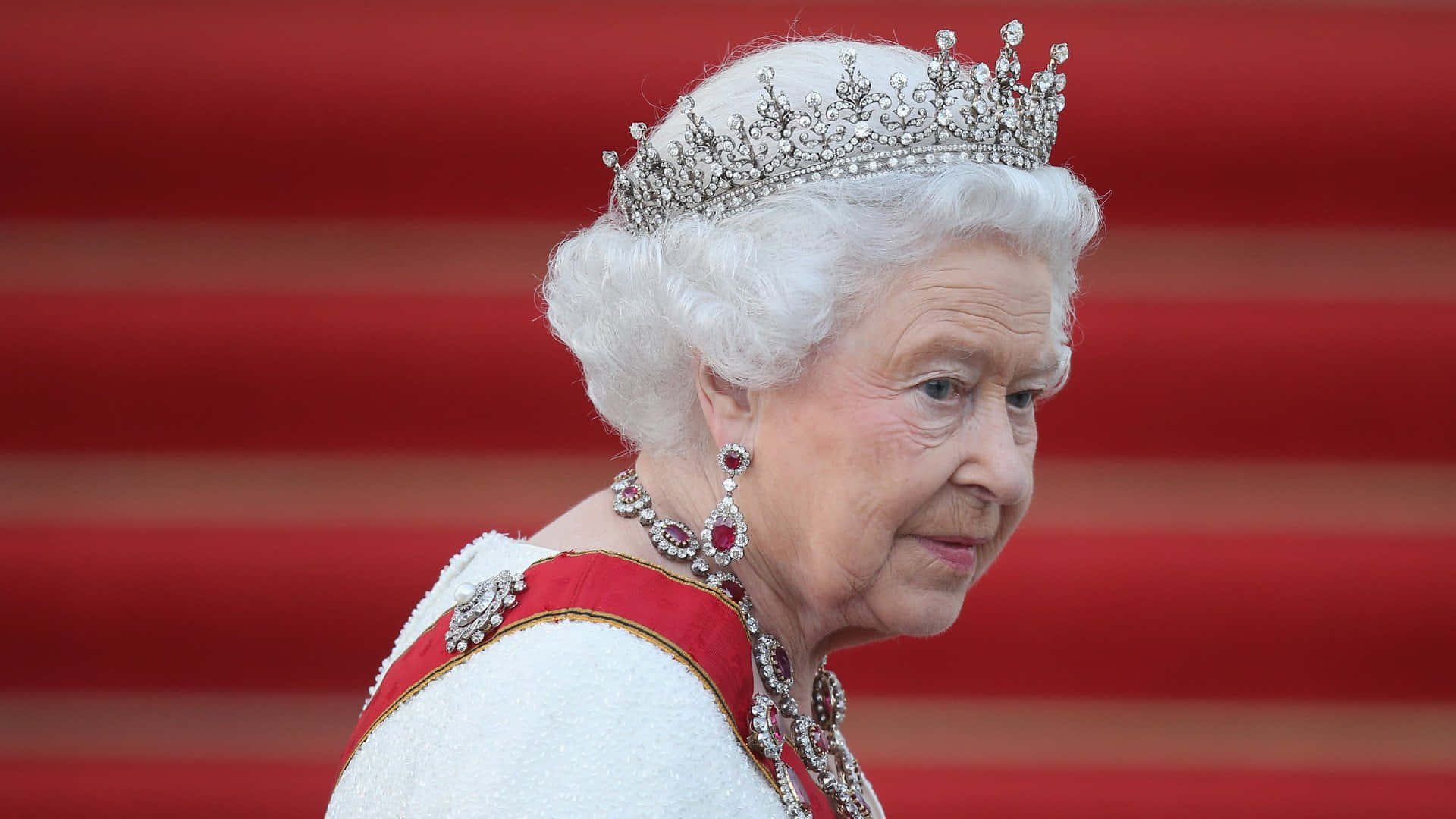Queen Elizabeth 桌布 壁紙 — 伊利沙伯二世女王身穿華麗服飾，佩戴閃爍鑽石冠冕和紅寶石耳環，背景是鮮豔的紅地毯。