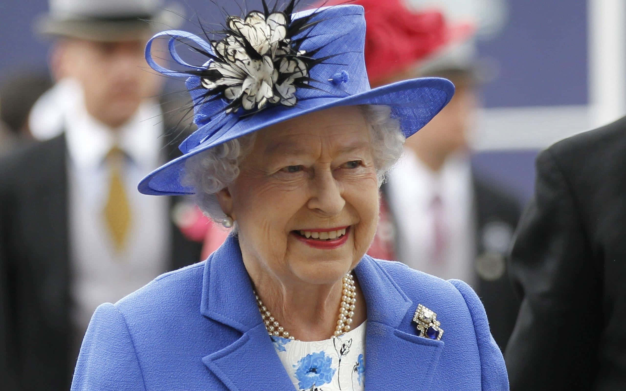Queen Elizabeth 桌布 壁紙 — 伊利沙伯二世女王身穿鮮藍色外套和配襯帽子，帽上飾有黑白羽毛，溫暖地微笑。