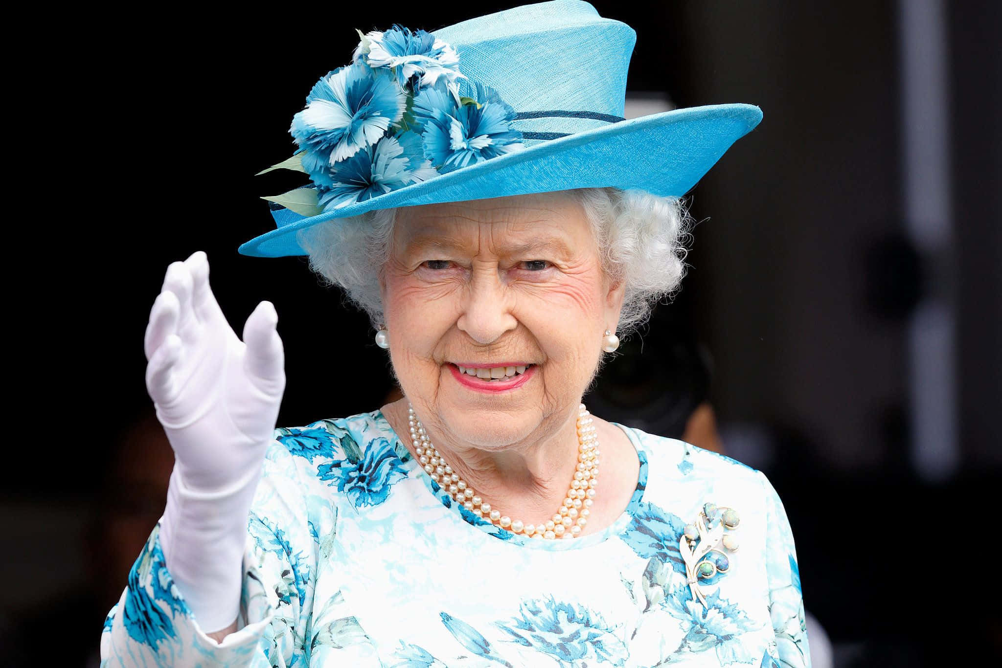 Queen Elizabeth 桌布 壁紙 — 伊利沙伯二世女王身穿鮮豔的藍色花卉套裝，頭戴飾有藍色花朵的配套帽子，溫暖地微笑揮手。