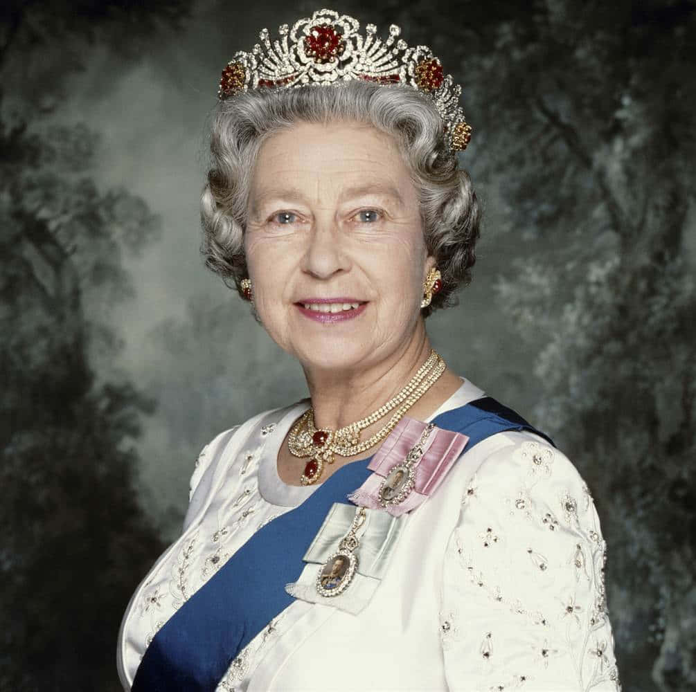 Queen Elizabeth 桌布 壁紙 — 伊利沙伯二世女王面帶微笑，威嚴地佩戴著鑲有珠寶的王冠、珍珠項鍊和藍色肩帶，背景是柔和的森林。