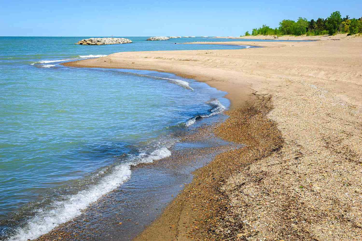 在清澈湛藍的天空下，海浪湧向卵石海灘，遠處有樹木。