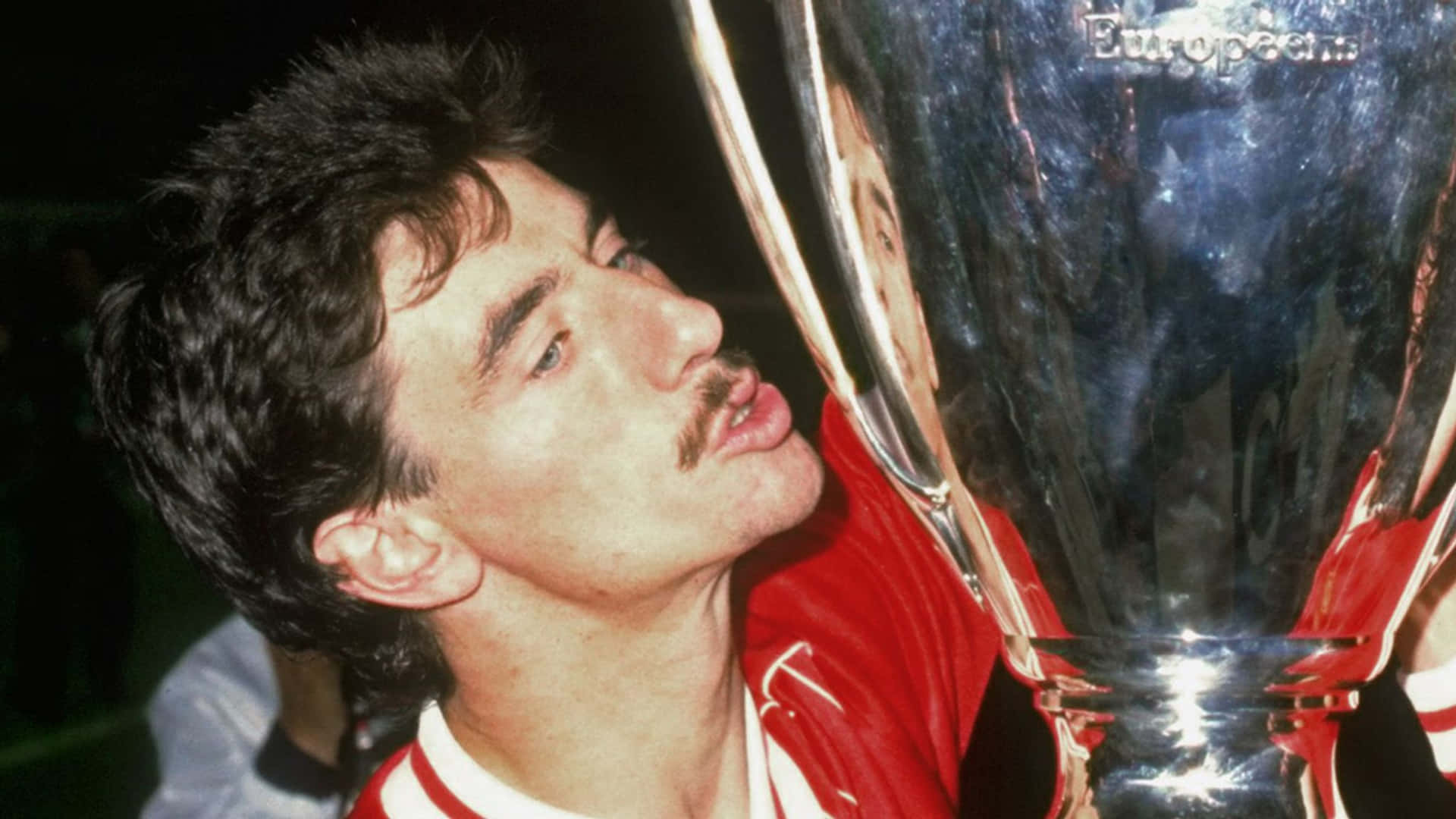 伊恩·魯什1984年聯賽盃攝影 壁紙 — 一位身穿紅色球衣的勝利足球員，抬頭望著靠近他臉龐的閃亮銀色歐洲盃獎盃。