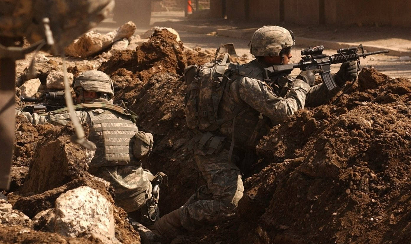 伊拉克戰爭士兵 壁紙 — 兩名身穿迷彩裝備的士兵在塵土飛揚的戰場上躲在土堆後面瞄準步槍。