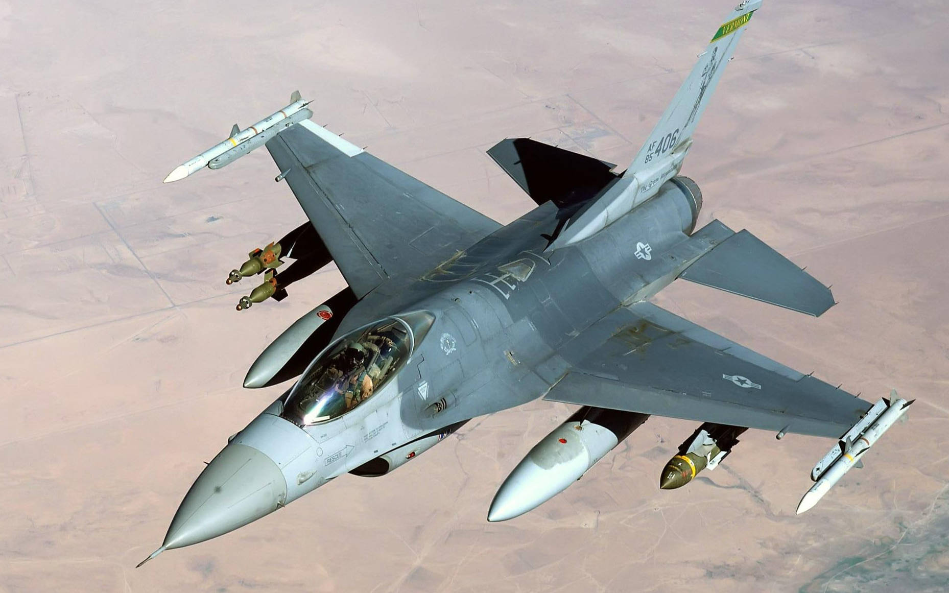 伊拉克沙漠F-16戰鬥機 壁紙 — 一架灰色戰鬥機在沙漠景觀上空翱翔，機翼下攜帶導彈和炸彈。