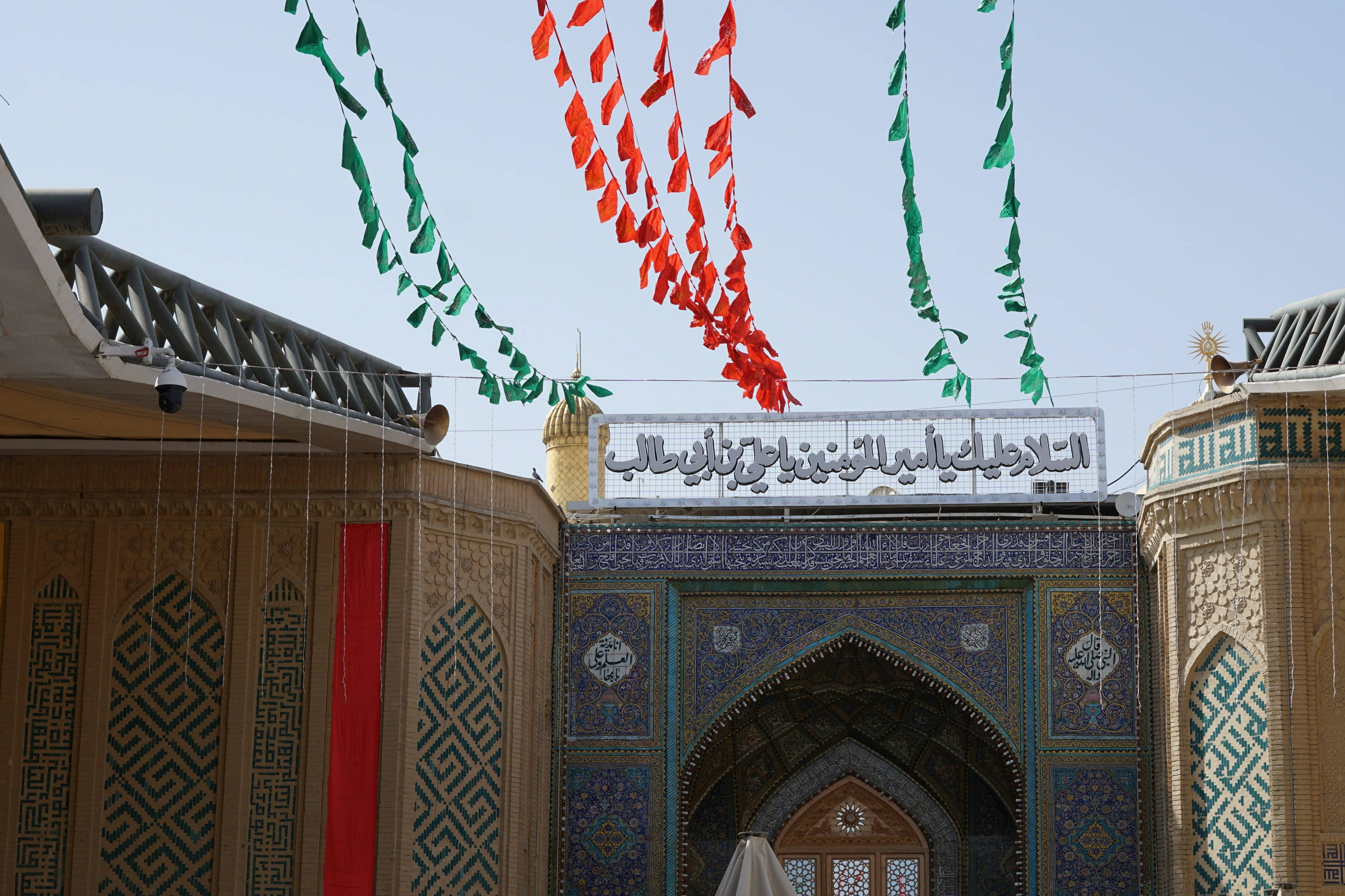 伊拉克納傑夫伊瑪目阿里聖殿 壁紙 — 一座宏偉的伊斯蘭清真寺入口，飾有精緻的藍色瓷磚，紅綠旗幟在晴朗的天空下高高懸掛。