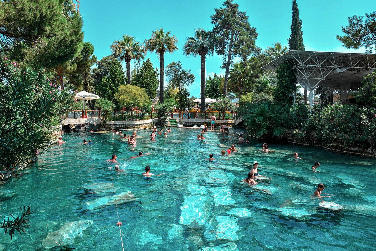 伊斯坦堡人工建造的克麗奧佩特拉池 壁紙 — 人們在清澈的藍天下，在茂密的棕櫚樹和綠樹環繞的充滿活力的綠松石色戶外游泳池中游泳和放鬆。