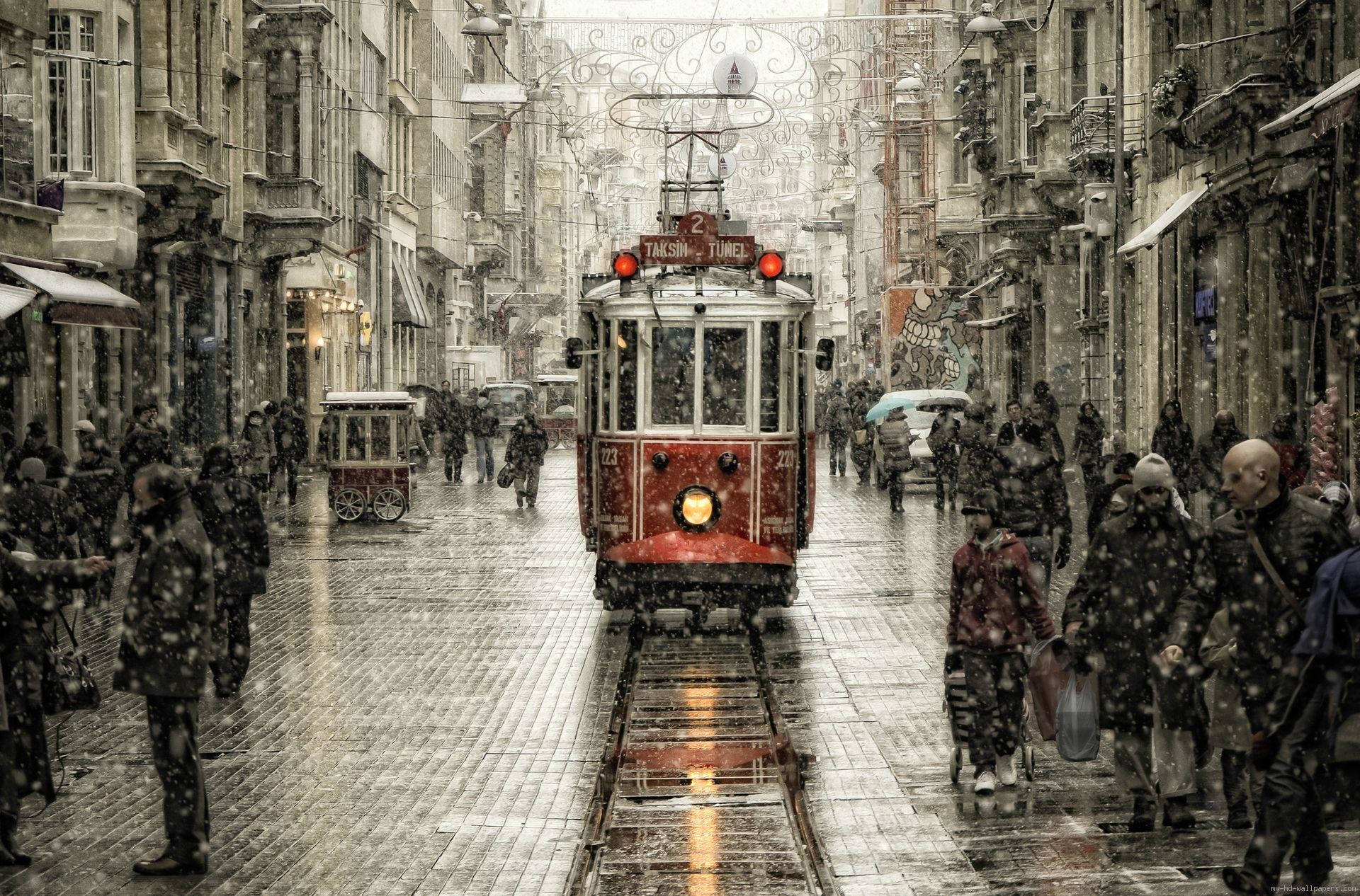 伊斯坦堡著名的鐵路線 壁紙 — 一輛紅色歷史電車在積雪的歐洲街道上滑行，行人則在飄雪中行走在濕滑的鵝卵石路上。