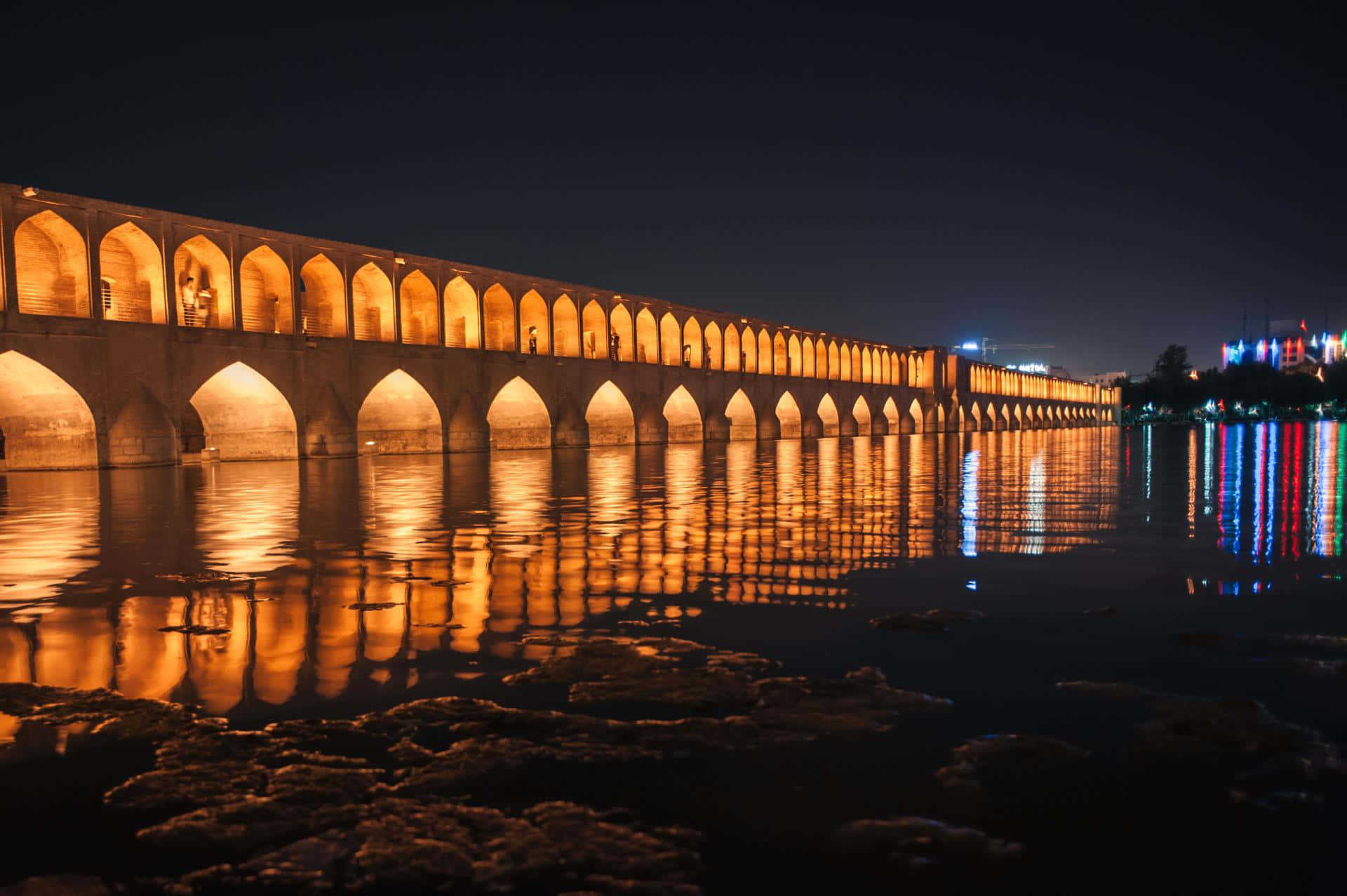 伊斯法罕阿拉維迪汗橋夜景燈光 壁紙 — 一座歷史悠久的石橋，發光的拱門在漆黑的夜空下，溫暖地倒映在平靜的水面上。