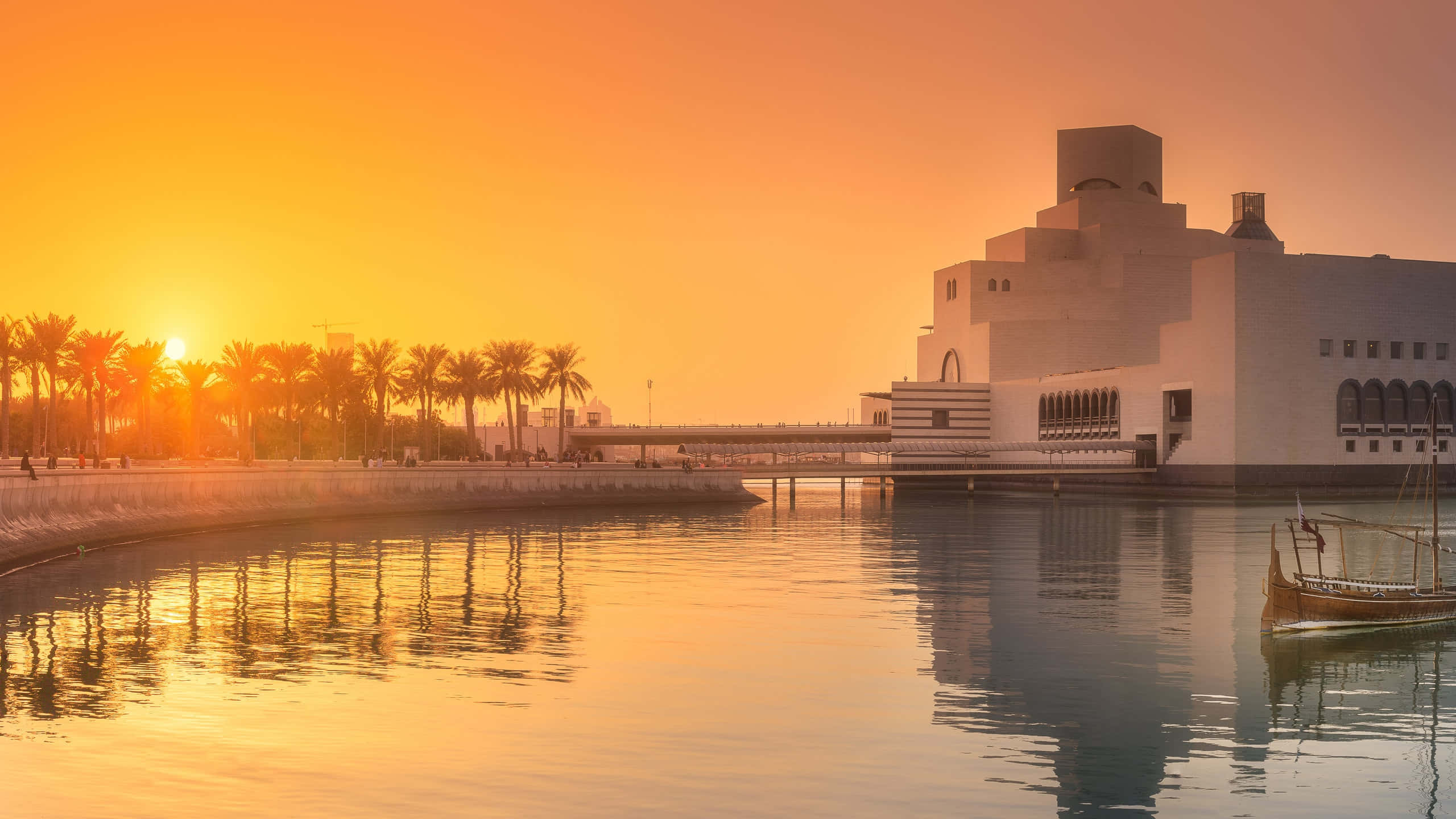 伊斯蘭藝術博物館在夕陽下 壁紙 — 一座現代白色博物館建築在金色日落時分倒映在平靜的水面上，附近有棕櫚樹和傳統的單桅帆船。