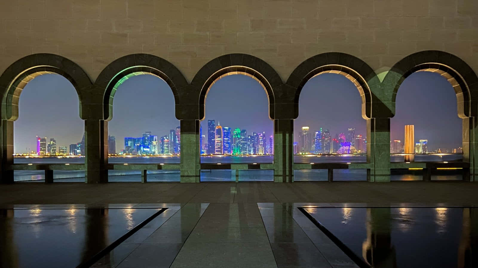 伊斯蘭藝術博物館拱門框住城市夜景 壁紙 — 夜晚景色，一排拱形石窗框住充滿活力的城市天際線，倒映在濕漉漉的路面上。