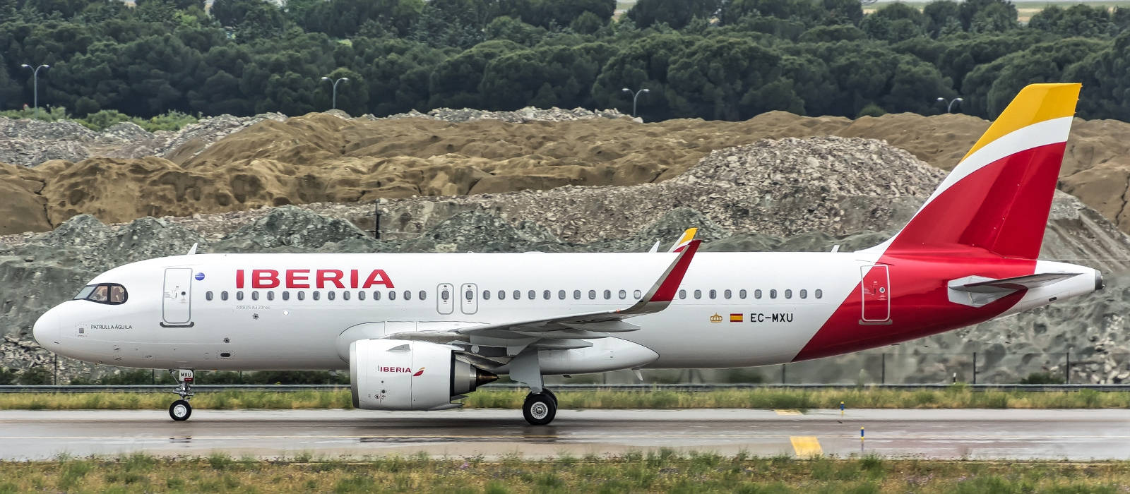 伊比利亞航空飛機在潮濕跑道上，附近有土色山丘和綠樹。
