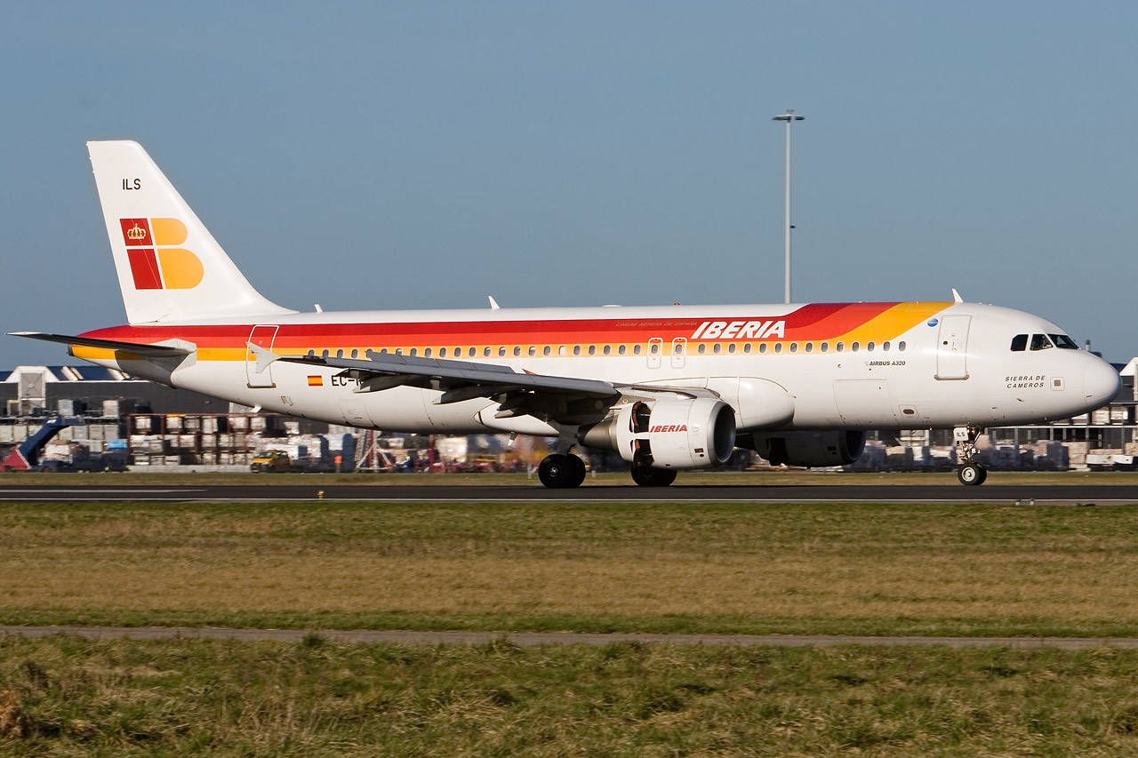 伊比利亞航空飛機在機場跑道滑行，後面有貨運集裝箱和建築物。