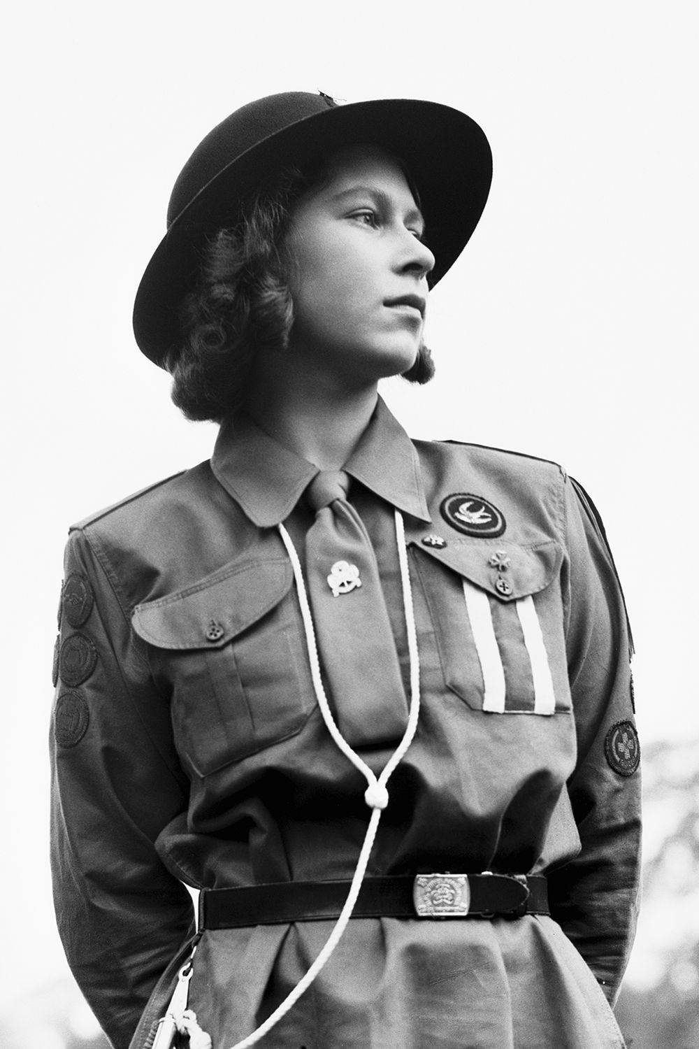 伊麗莎白女王 穿軍裝 壁紙 — 一位身穿復古童軍制服、戴著帽子和繩索領巾的年輕女性，在明亮的天空下向上凝視。