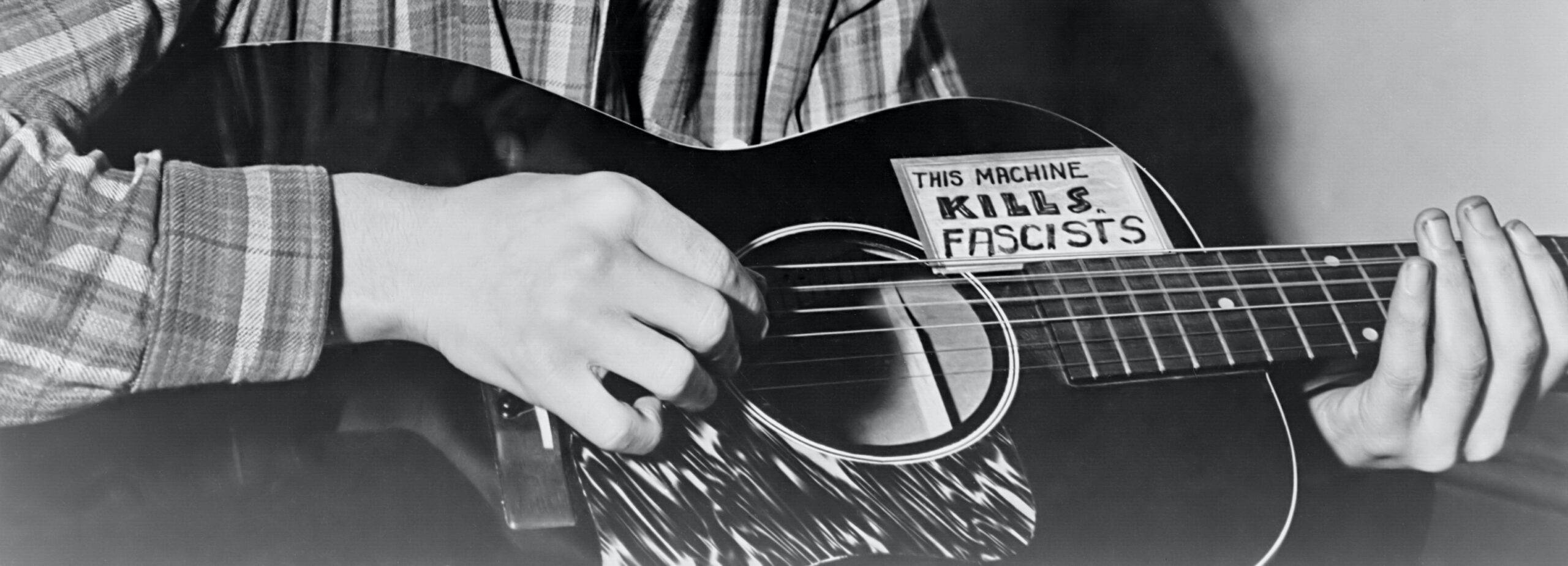 伍迪·蓋瑟瑞的吉他 壁紙 — 一個人彈奏木結他，結他身上貼有寫著「THIS MACHINE KILLS FASCISTS」的大膽貼紙。