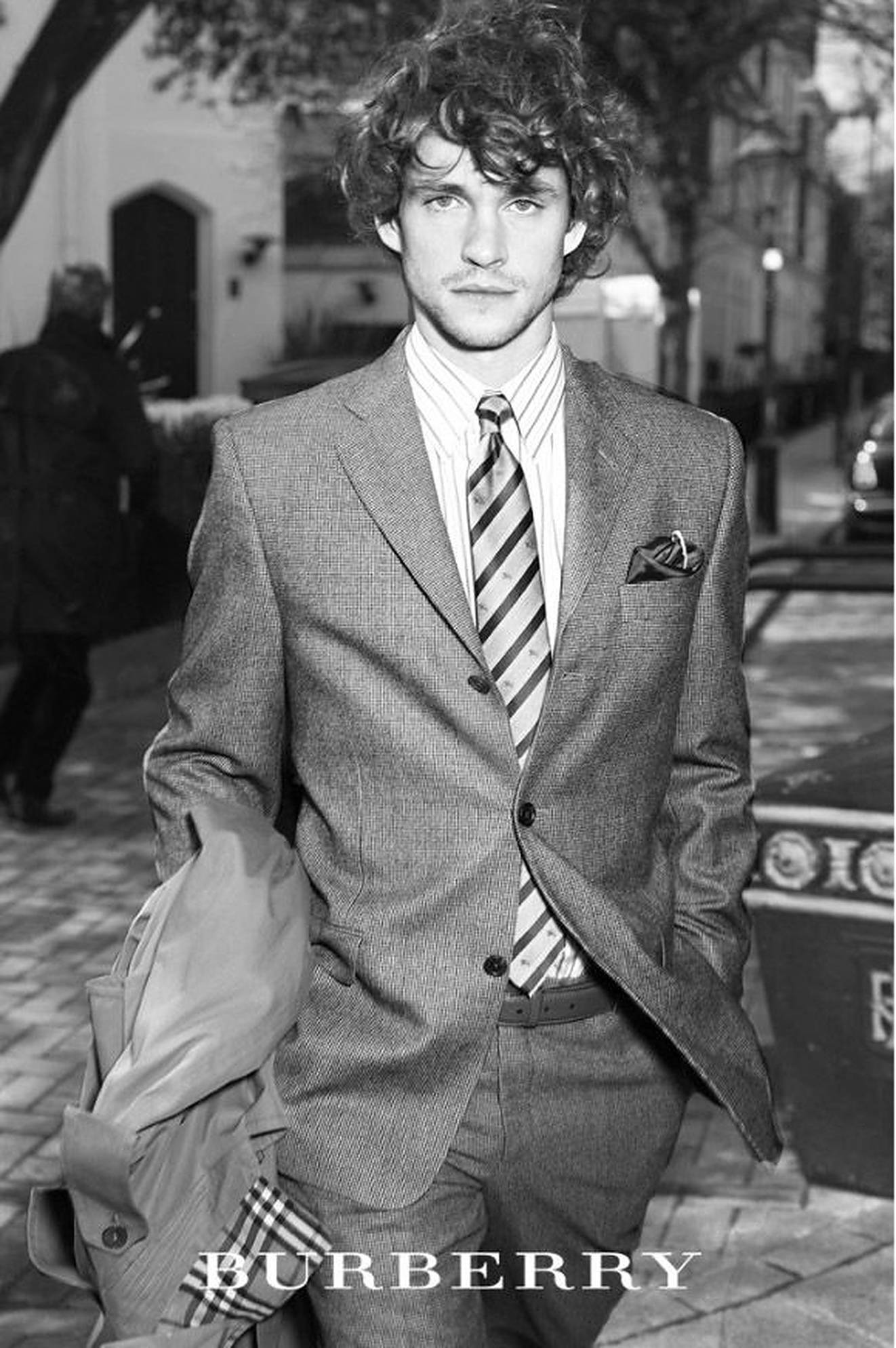 休·丹西身著 Burberry 壁紙 — 一位時尚男士穿著質感西裝和條紋領帶，自信地站在鵝卵石街道上，黑白風格。