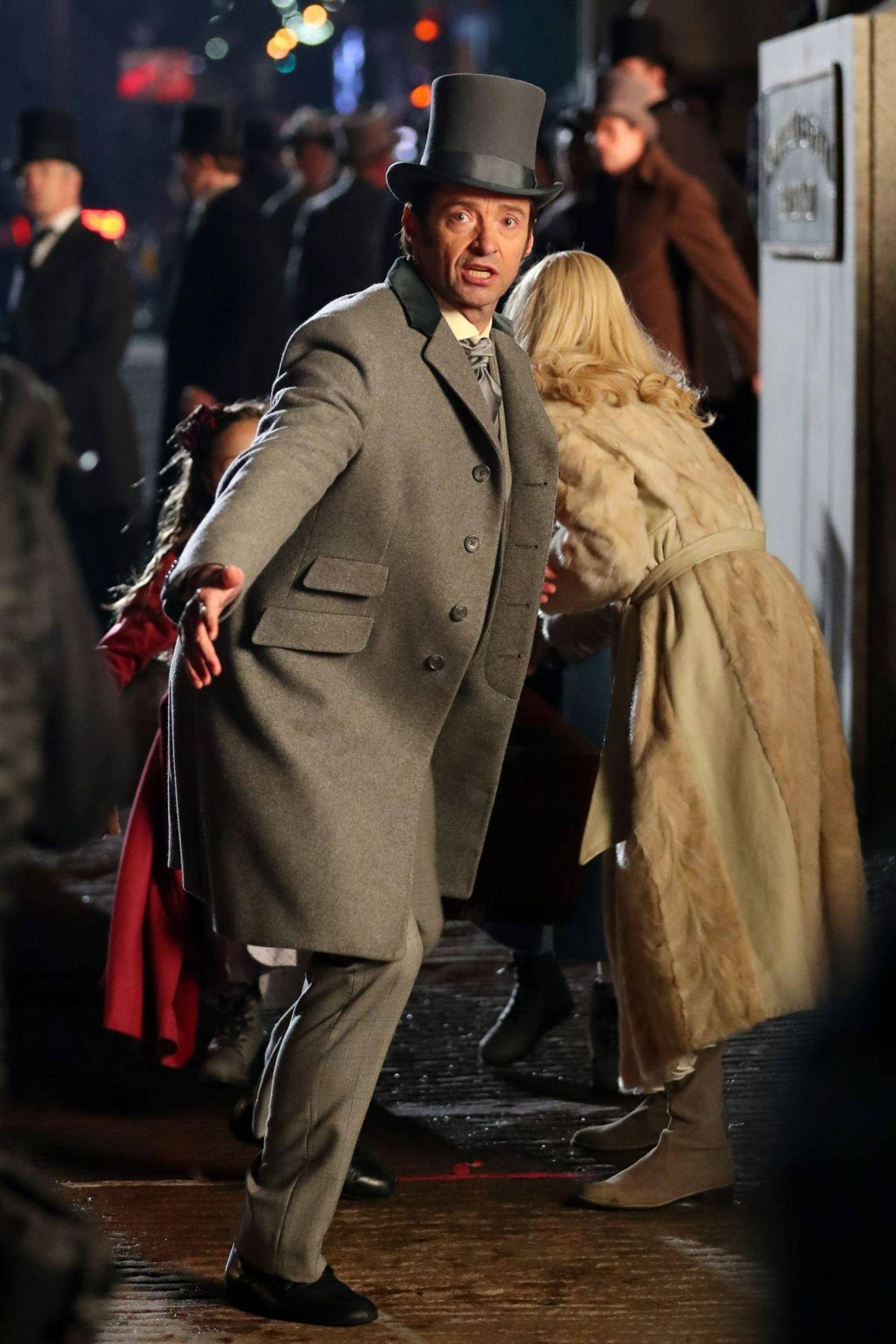 休·傑克曼在《 Greatest Showman 》 壁紙 — 一個戴灰色高帽、穿灰色大衣的男人在雨夜的城市街道上向前猛衝，而一個穿米色大衣的女人正在逃跑。