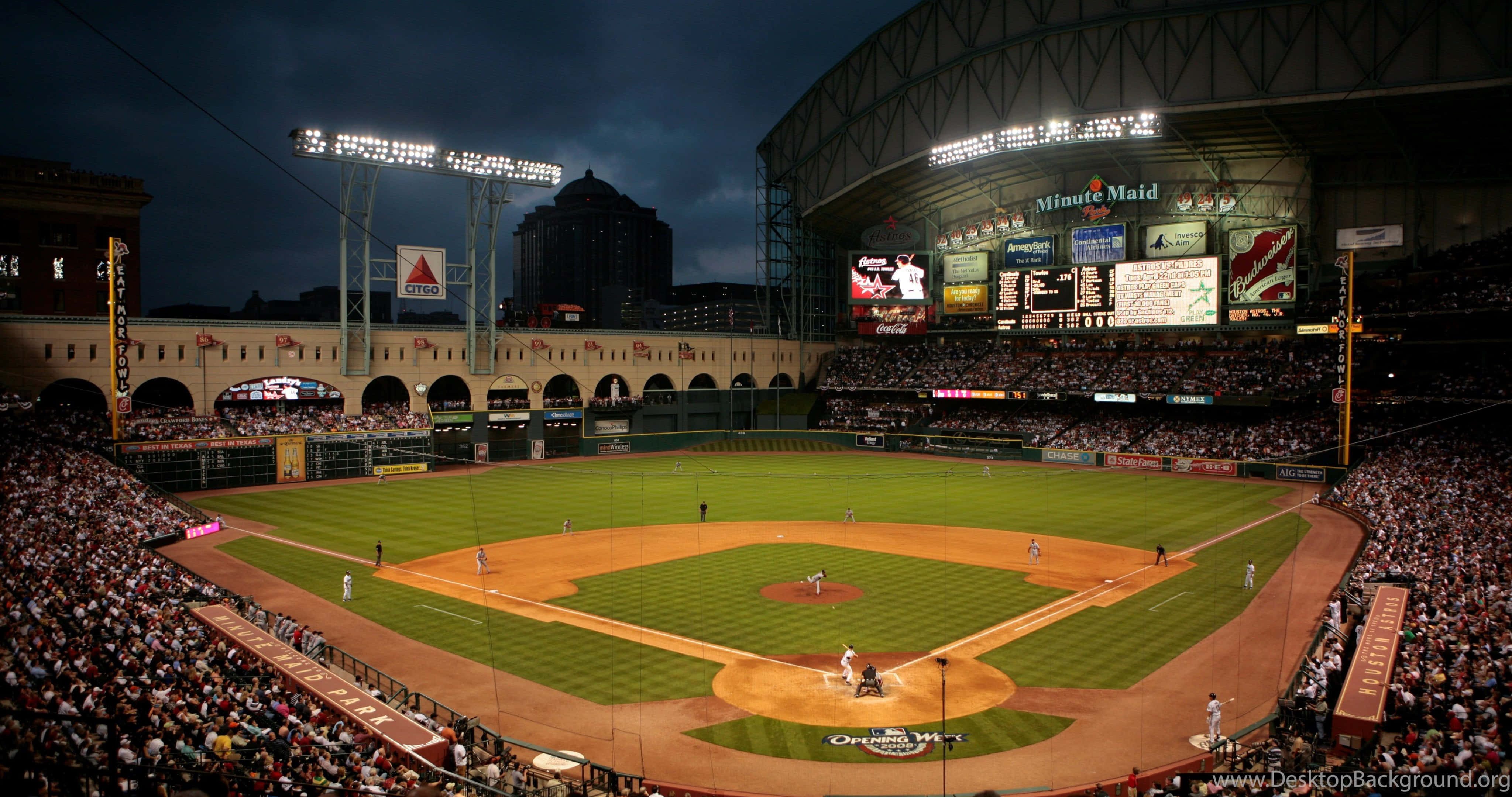 休士頓 Minute Maid Park 棒球場景觀背景 壁紙 — 黃昏時分擠滿人的棒球場，場上有球員，明亮的燈光照亮綠色草地和泥土內野。