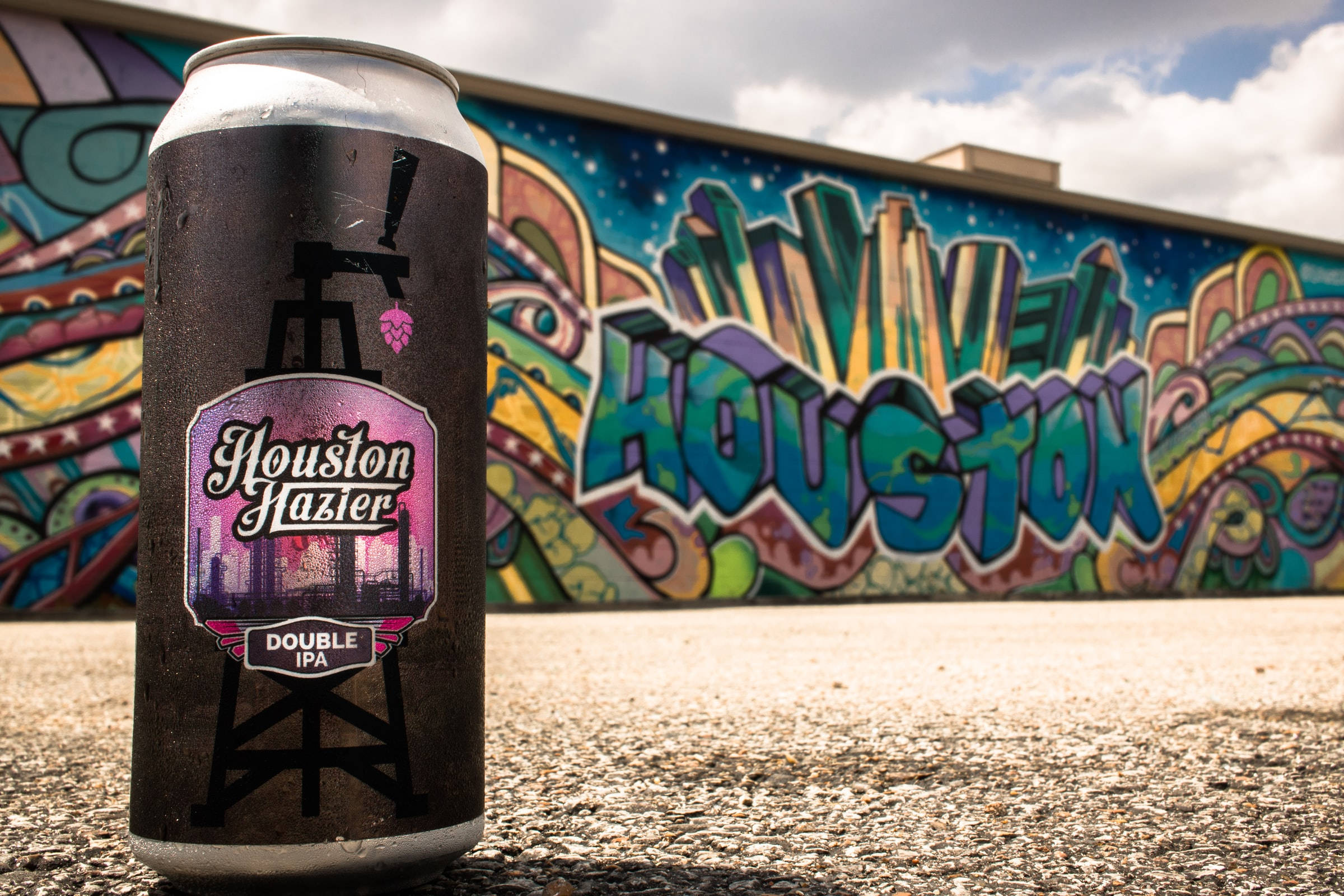 休斯頓塗鴉藝術風景 壁紙 — 一罐冰凍的Houston Hazier Double IPA啤酒放在人行道上，前面是充滿活力的塗鴉壁畫，在陰天天空下拼出Houston。
