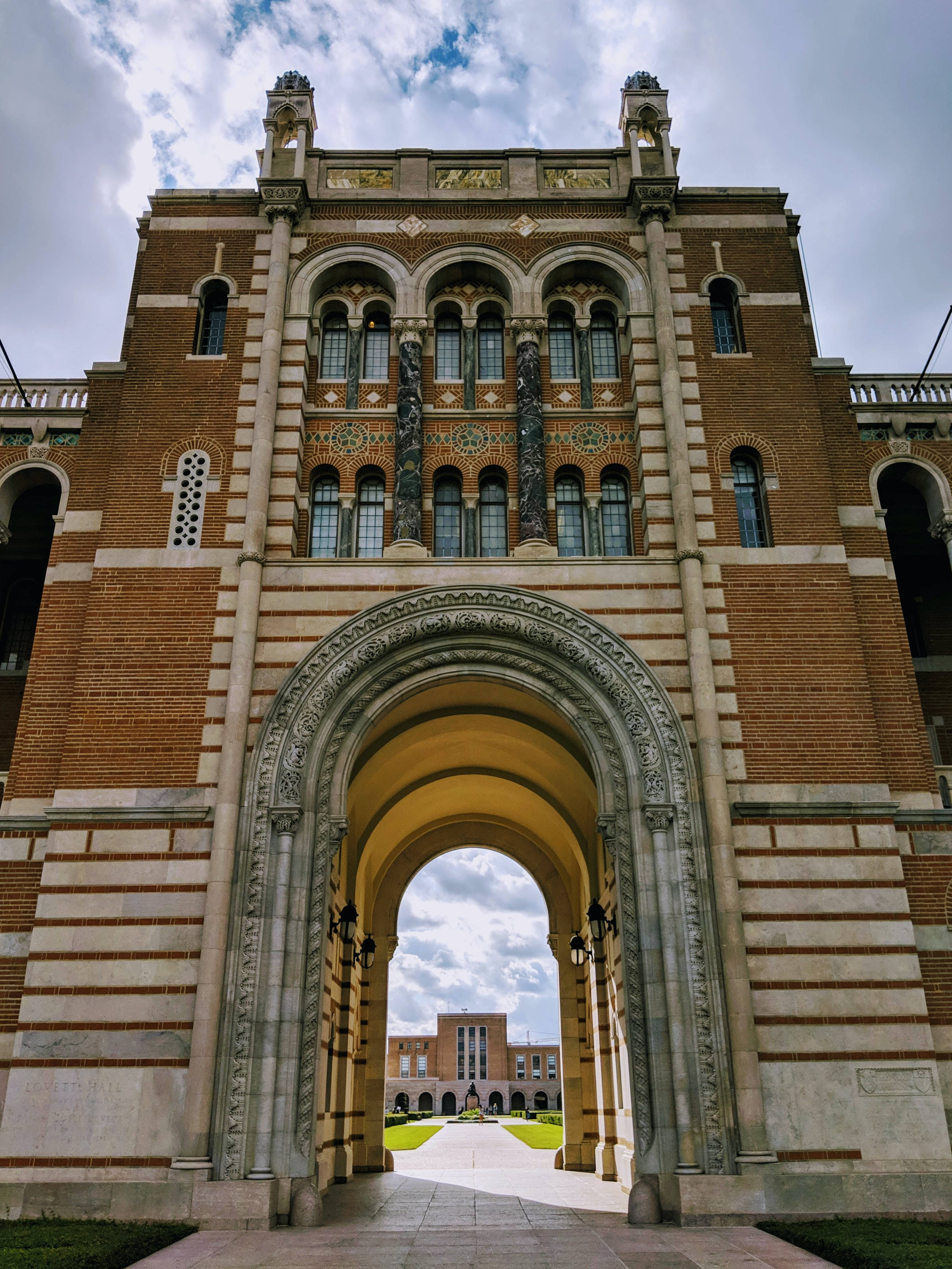 休斯頓萊斯大學壯麗校園風景 壁紙 — 一座宏偉的歷史磚砌建築，擁有華麗的拱門和精細的石雕，天空部分多雲。