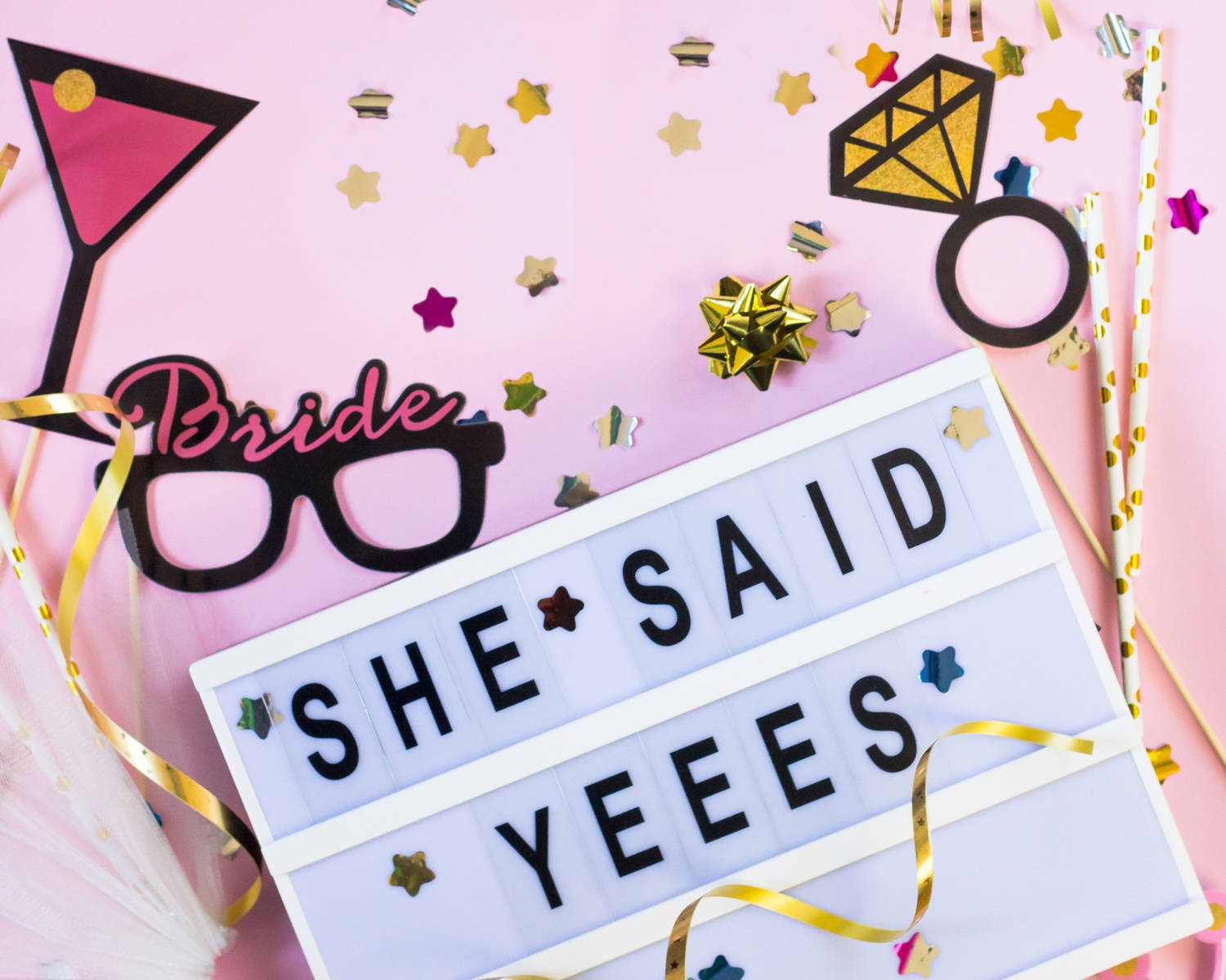 在粉紅色背景上，以寫著 'SHE SAID YEEES' 的燈箱和派對道具慶祝準新娘。