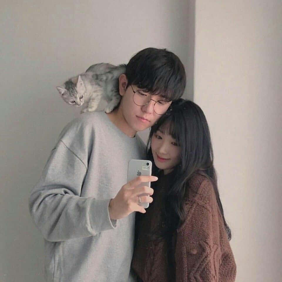 伴彼此穿越人生高低起伏 壁紙 — 一對年輕情侶在室內自拍，一隻灰色貓咪棲息在男士的肩膀上，背景是素牆。