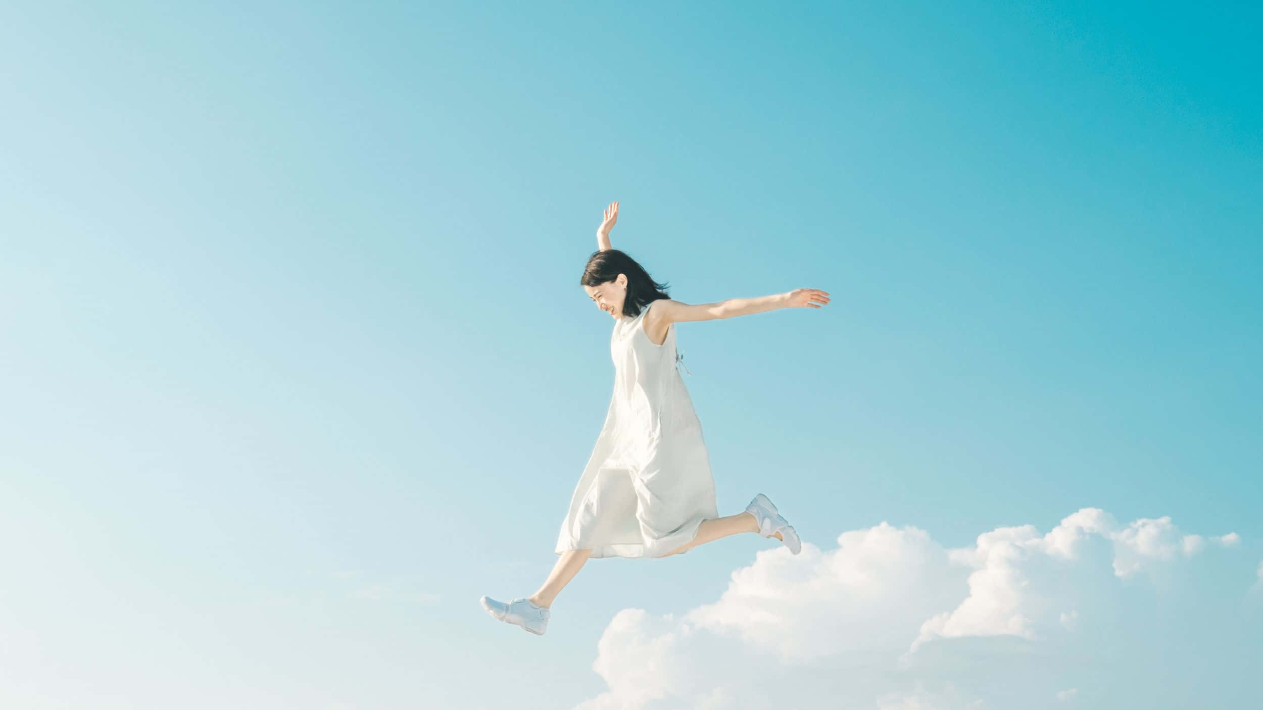 伸手摘星 壁紙 — 一位身穿白色連衣裙的女性在清澈的藍天和下方蓬鬆的雲朵中歡快地跳躍。