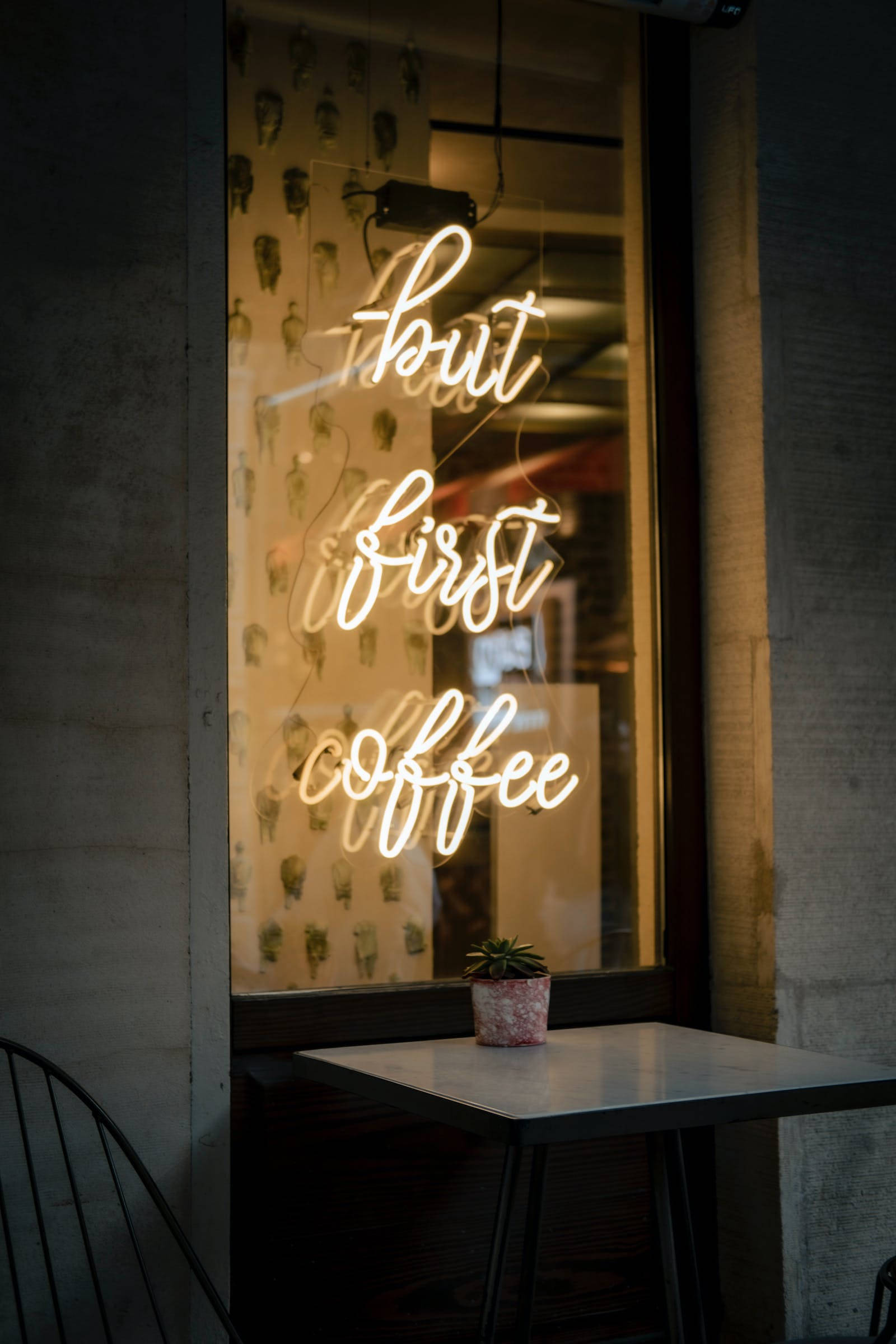 但先喝咖啡霓虹 iPhone 壁紙 — 一個發光的霓虹燈牌寫著「but first coffee」，照亮了夜間舒適的咖啡館窗戶，外面桌子上放著一盆小盆栽。