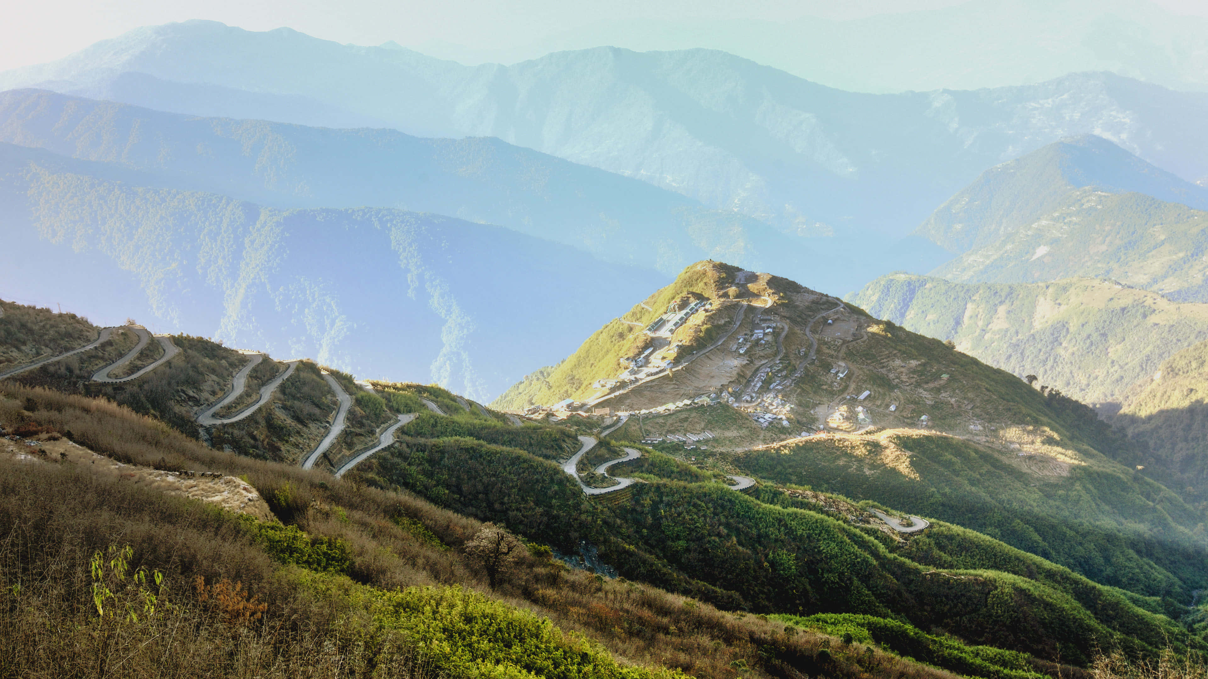 佈滿樹木的山間道路 壁紙 — 蜿蜒的山路在陽光照耀的綠色山丘間蜿蜒，層層疊疊的藍色山峰逐漸消失在朦朧的遠方。