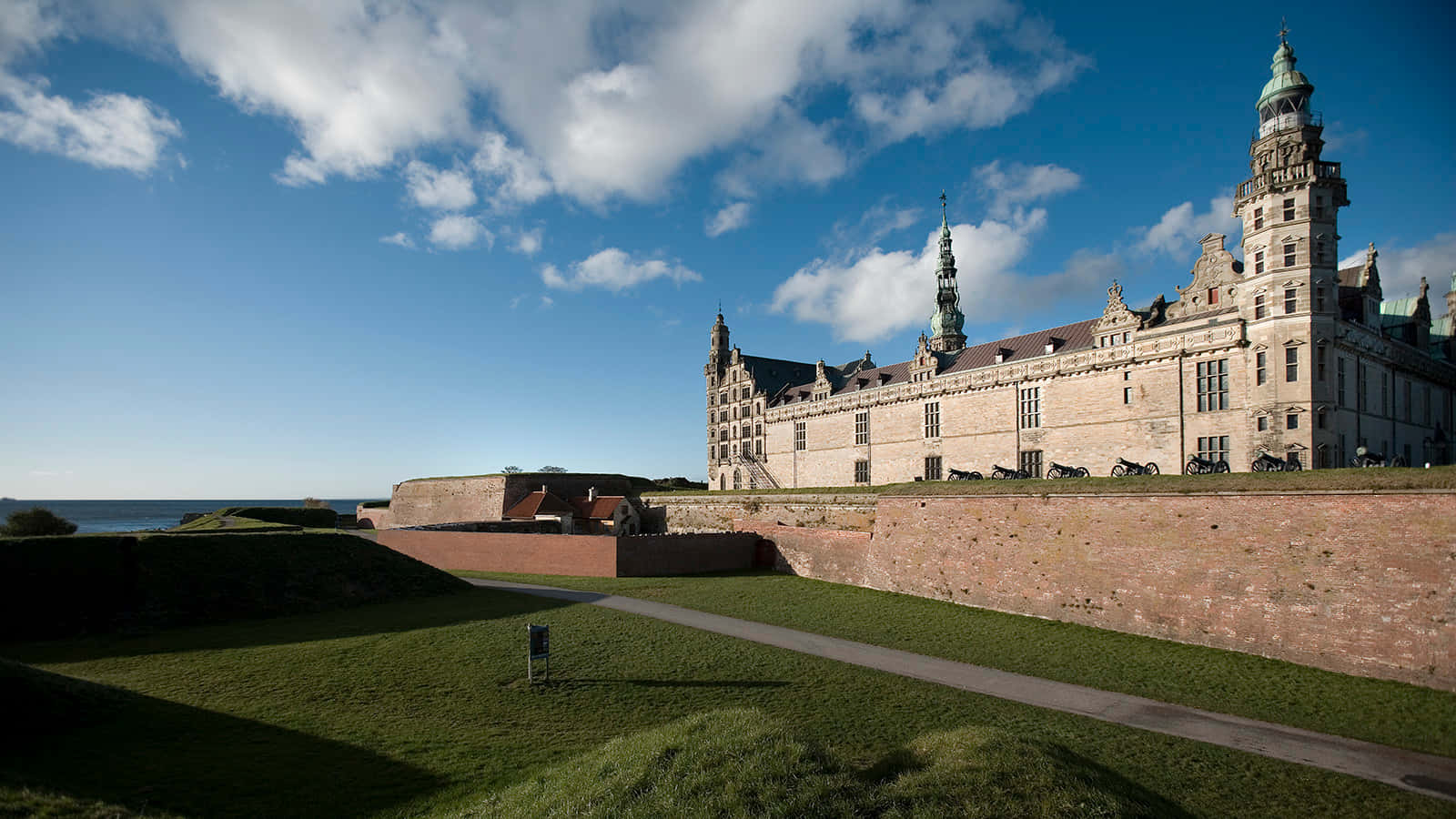 一座大型石造城堡，帶有塔樓和尖頂，位於青草覆蓋的山丘上，天空湛藍，白雲飄飄。