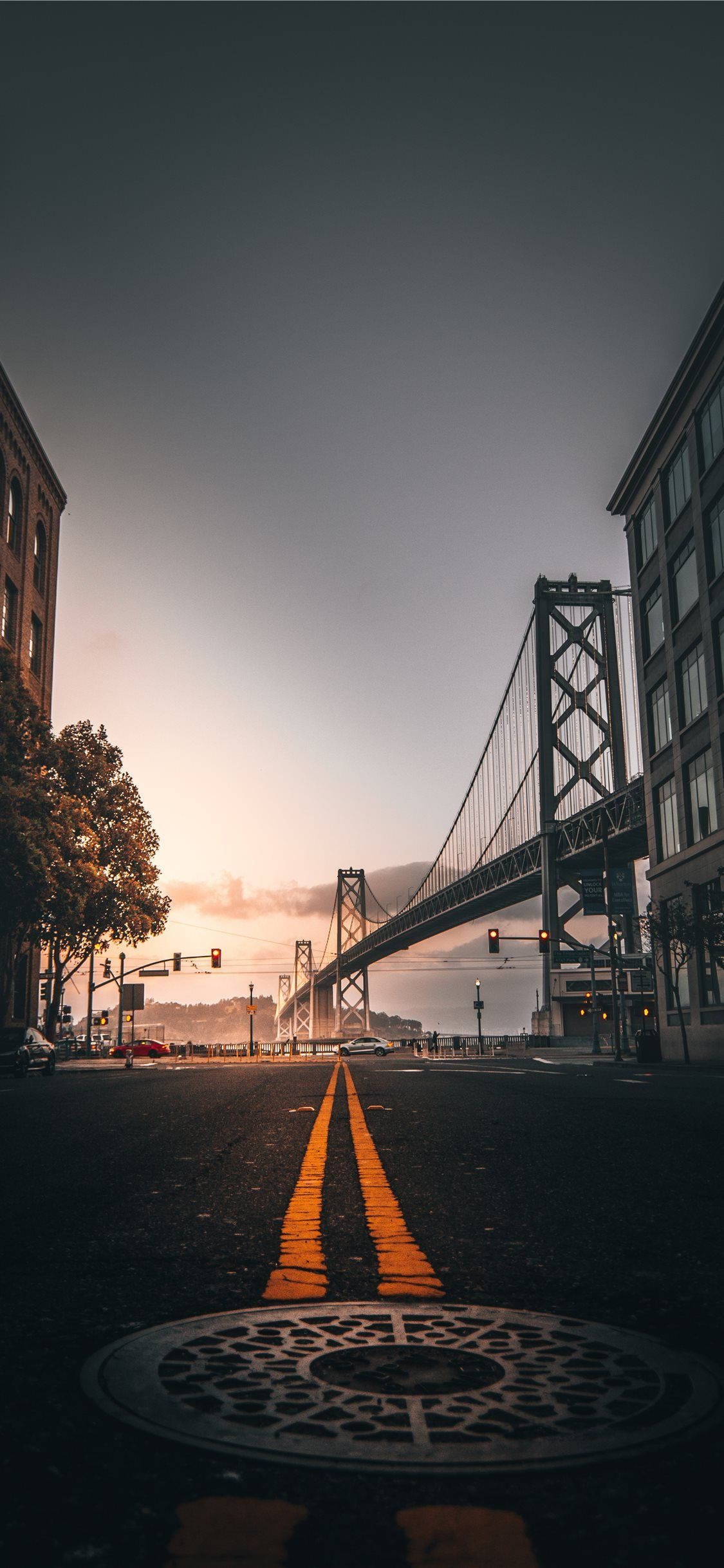 San Francisco Phone 桌布 壁紙 — 低角度城市街道景觀，通往日落時分的吊橋，瀝青路上有溫暖的金色線條，前景有裝飾性沙井蓋。