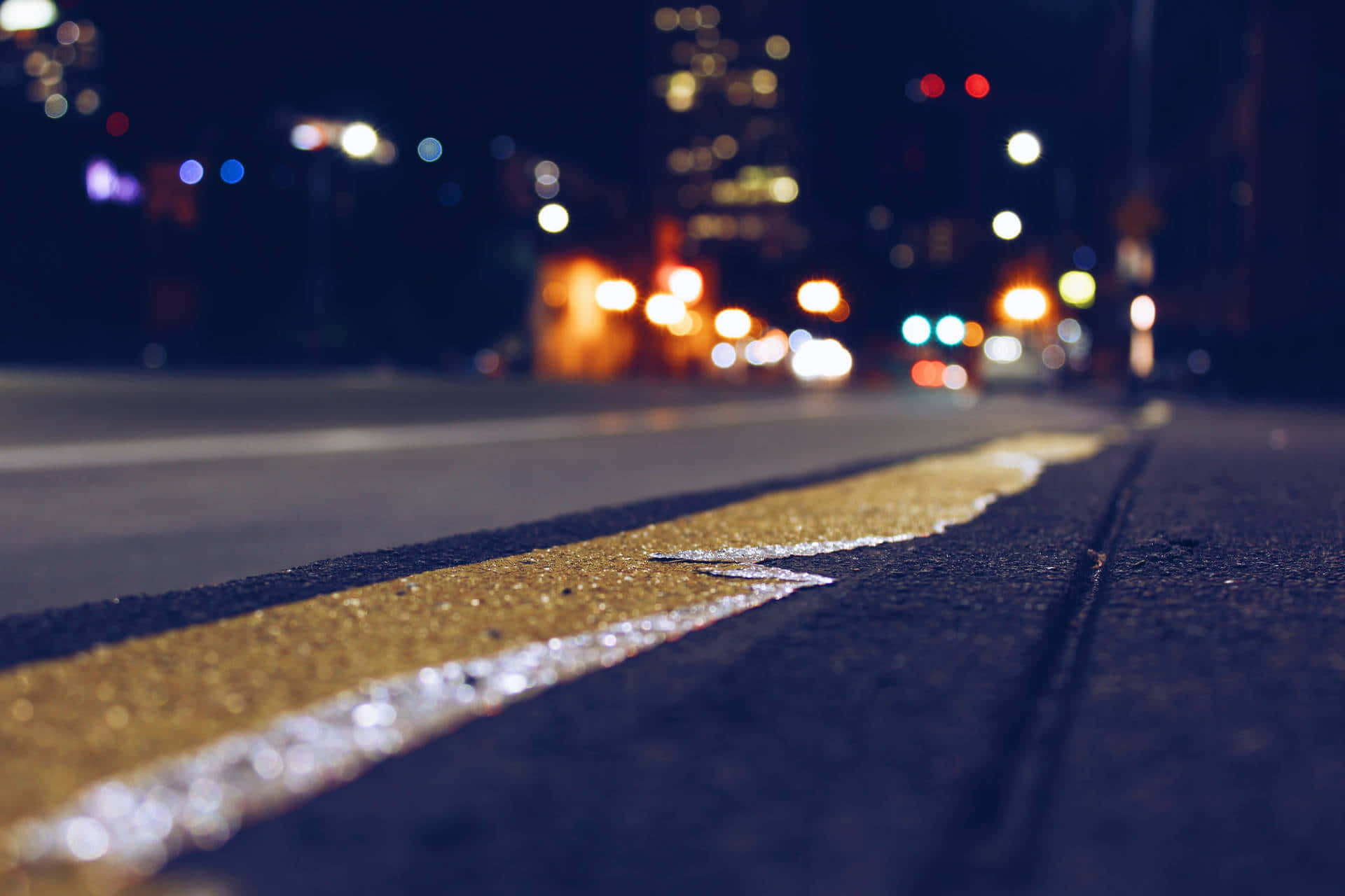 Dslr Blur 桌布 壁紙 — 低角度夜拍，濕漉漉的黃色道路標線在城市燈光下閃耀，背景是模糊的車流散景。