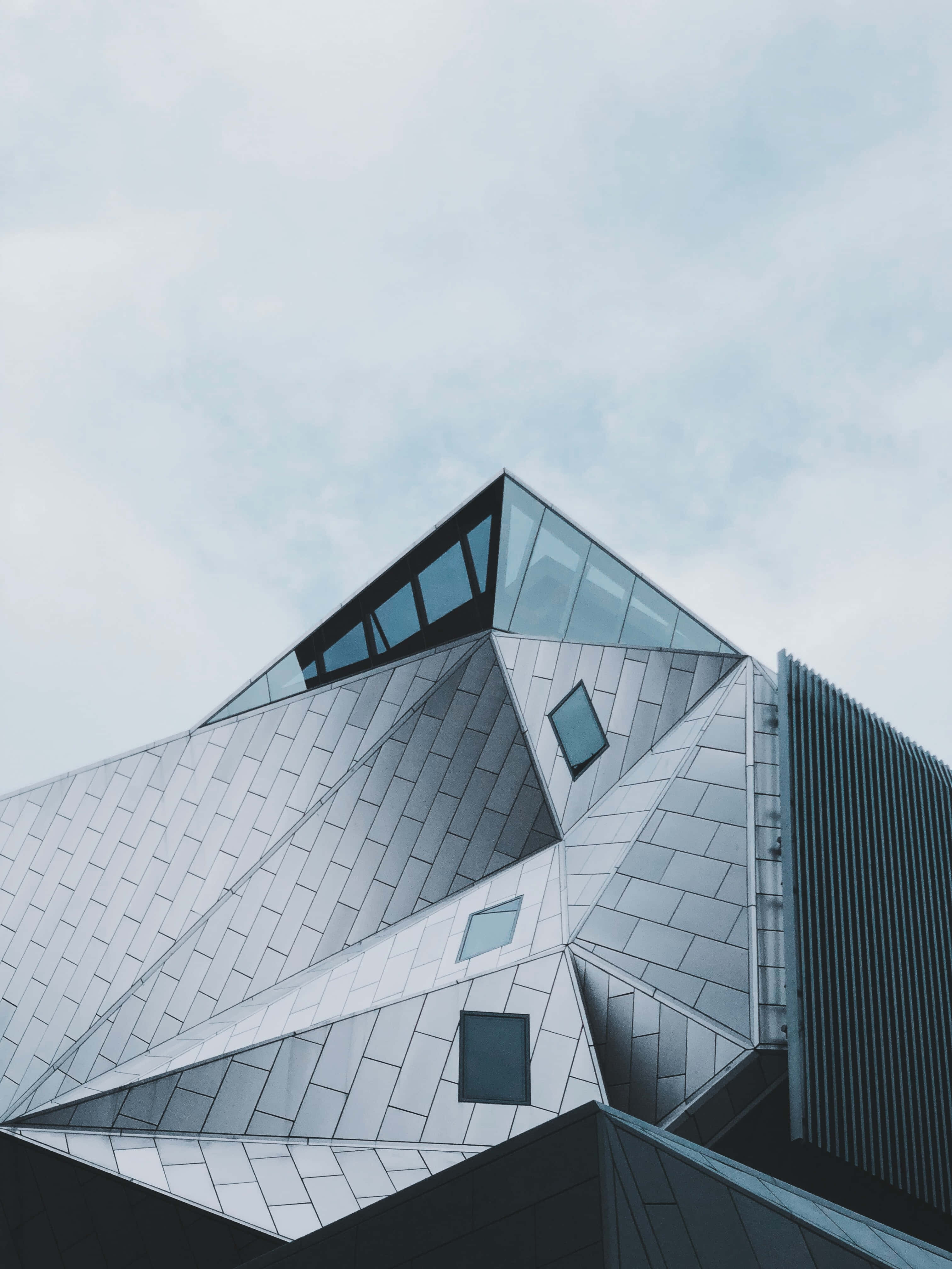 Modernism 桌布 壁紙 — 低角度拍攝一座現代幾何建築，配有銀色金屬面板和玻璃窗，背景是淡色天空。