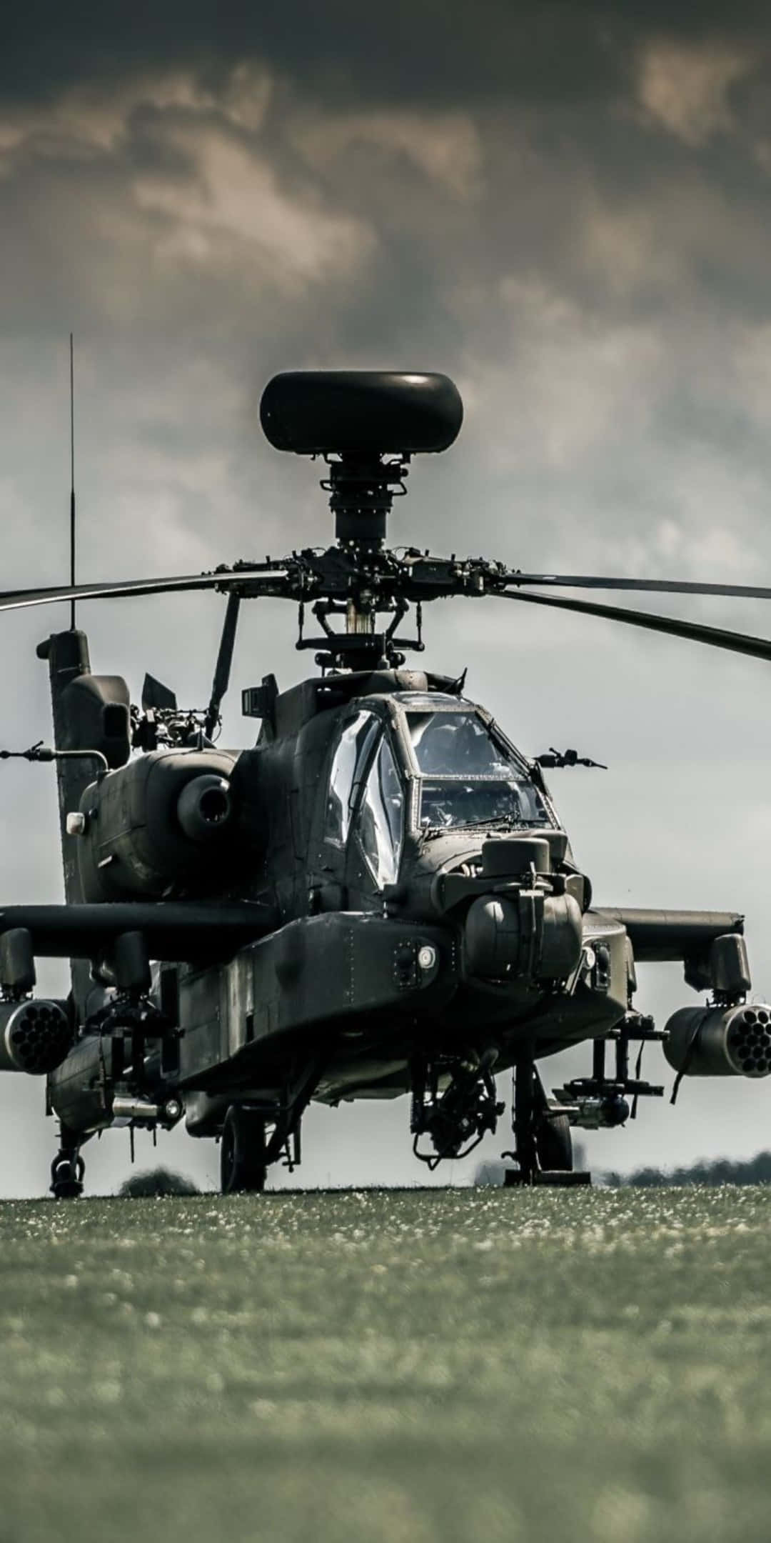 低角度拍攝的AH-64 Apache直升機 - Pixel 3背景 壁紙 — 一架軍用攻擊直升機在戲劇性的多雲天空下，佇立在草地上。