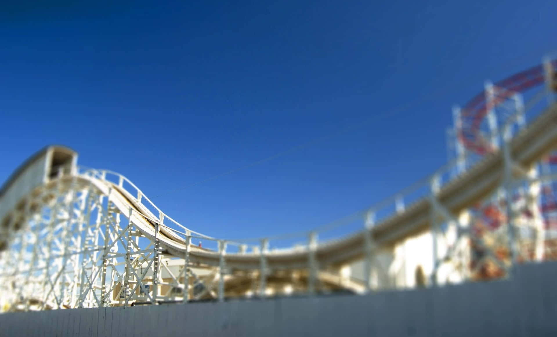 Roller Coaster 桌布 壁紙 — 低角度移軸視角下的白色木製過山車，背景是清澈的藍天，前景模糊。