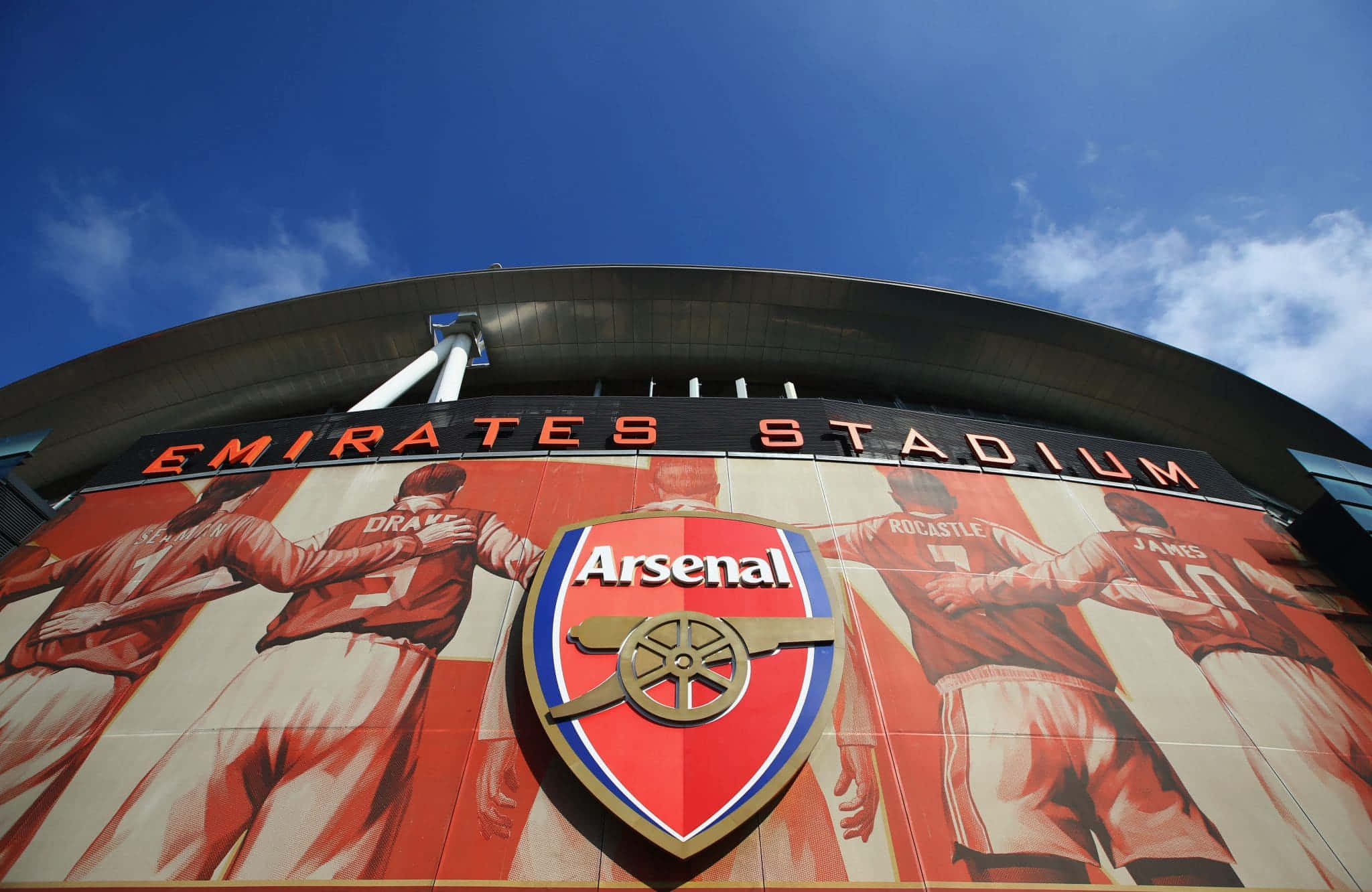 Arsenal 桌布 壁紙 — 低角度觀看酋長球場外牆，上有阿仙奴會徽及球員壁畫，背景為晴朗藍天。