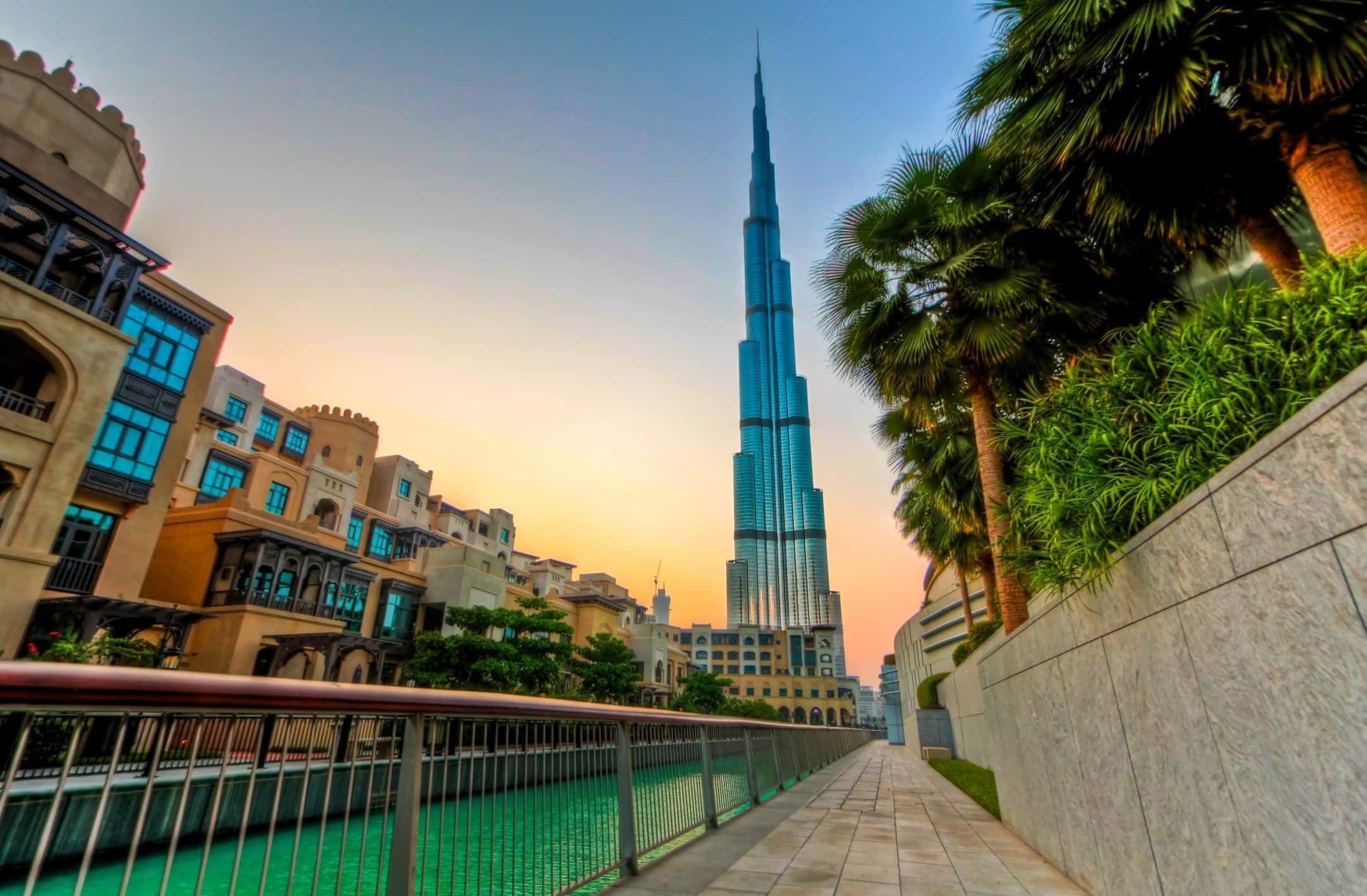 Burj Khalifa 桌布 壁紙 — 低角度觀賞哈利法塔在日落時分聳立於運河步道上，附近有棕櫚樹和傳統建築。