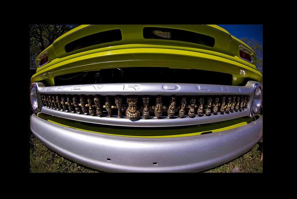 Car Grill 桌布 壁紙 — 低角度魚眼鏡頭拍攝捕捉到一輛復古青檸綠色雪佛蘭卡車的鍍鉻格柵，裝飾著部落面具，背景是晴朗的藍天。