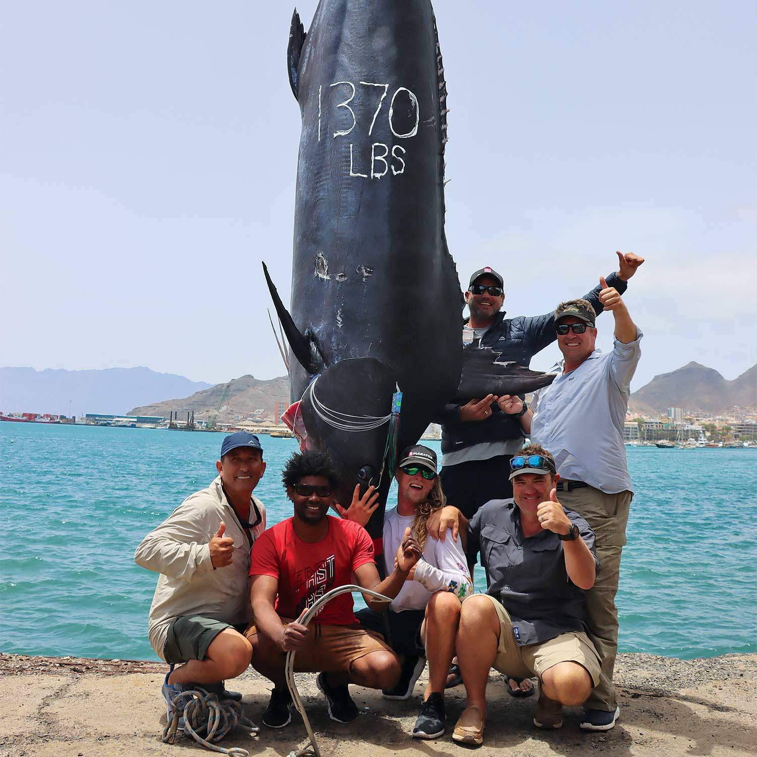 六位開心的釣魚人士在陽光明媚的碼頭上慶祝捕獲一條標示為1370磅的巨大吞拿魚。