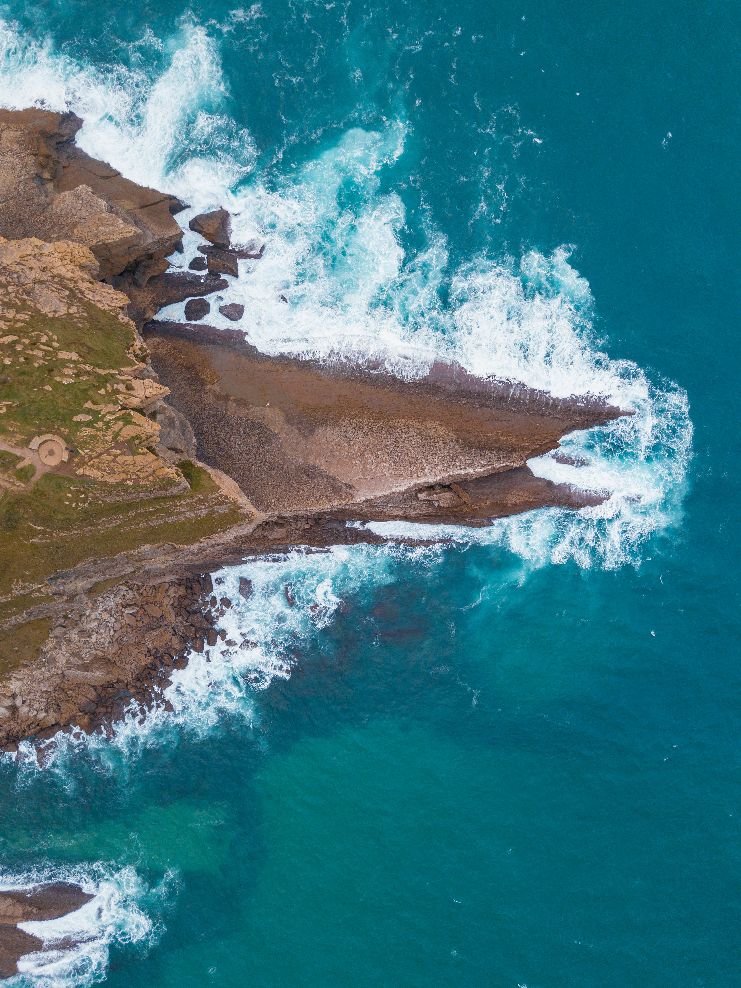 佛得角海景俯瞰 壁紙 — 從高空俯瞰，碧綠色的海浪拍打著崎嶇的懸崖，懸崖上有綠色草叢和一個小型圓形結構。