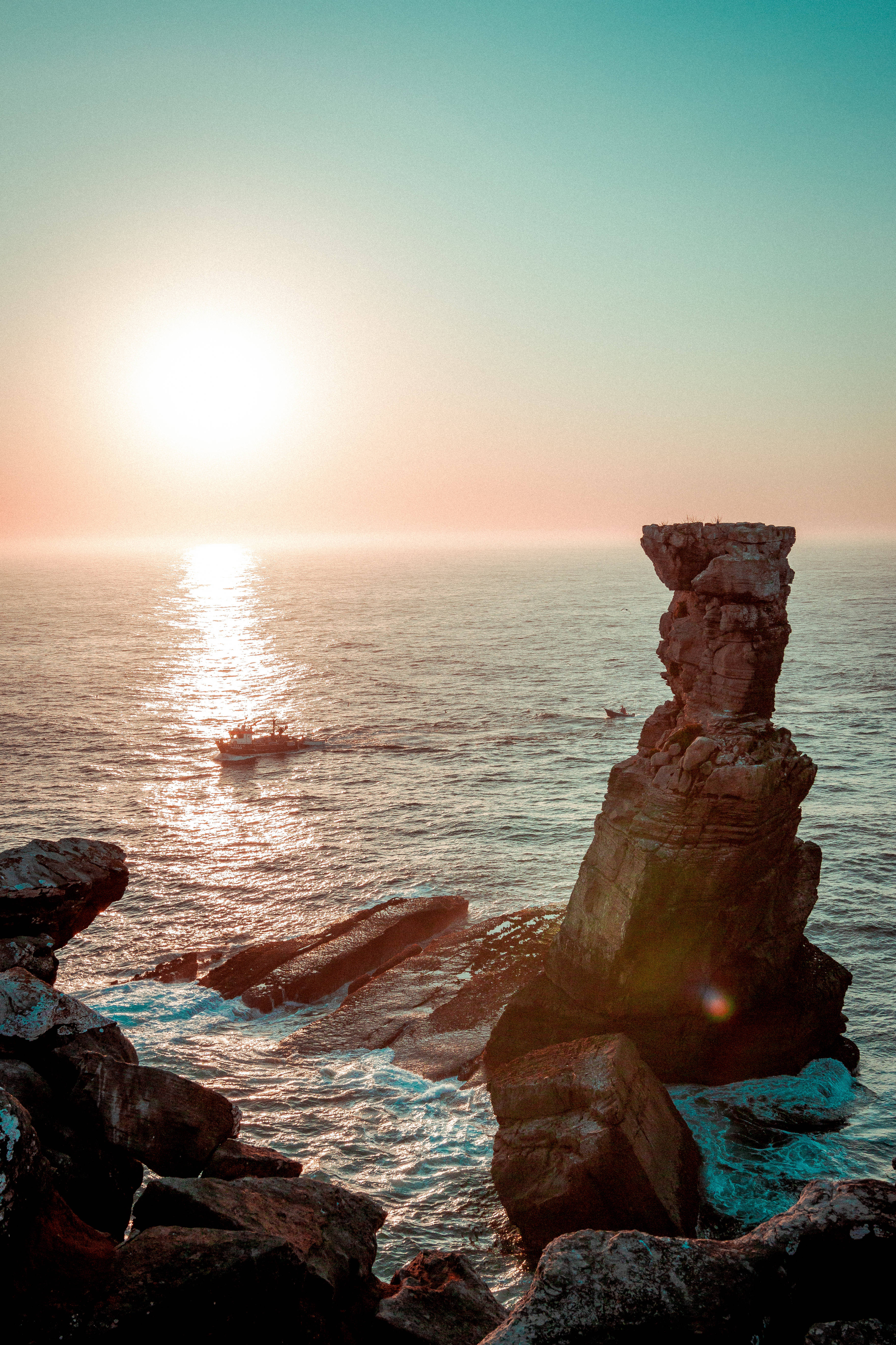 佛得角海洋與日落 壁紙 — 夕陽以溫暖的光芒沐浴著高聳的海蝕柱，一艘小船在平靜的海平線上滑行。