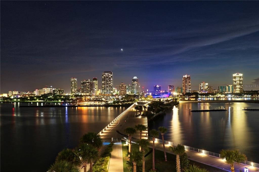 夜晚發光的城市天際線倒映在水中，有一條燈火通明的步道和棕櫚樹。