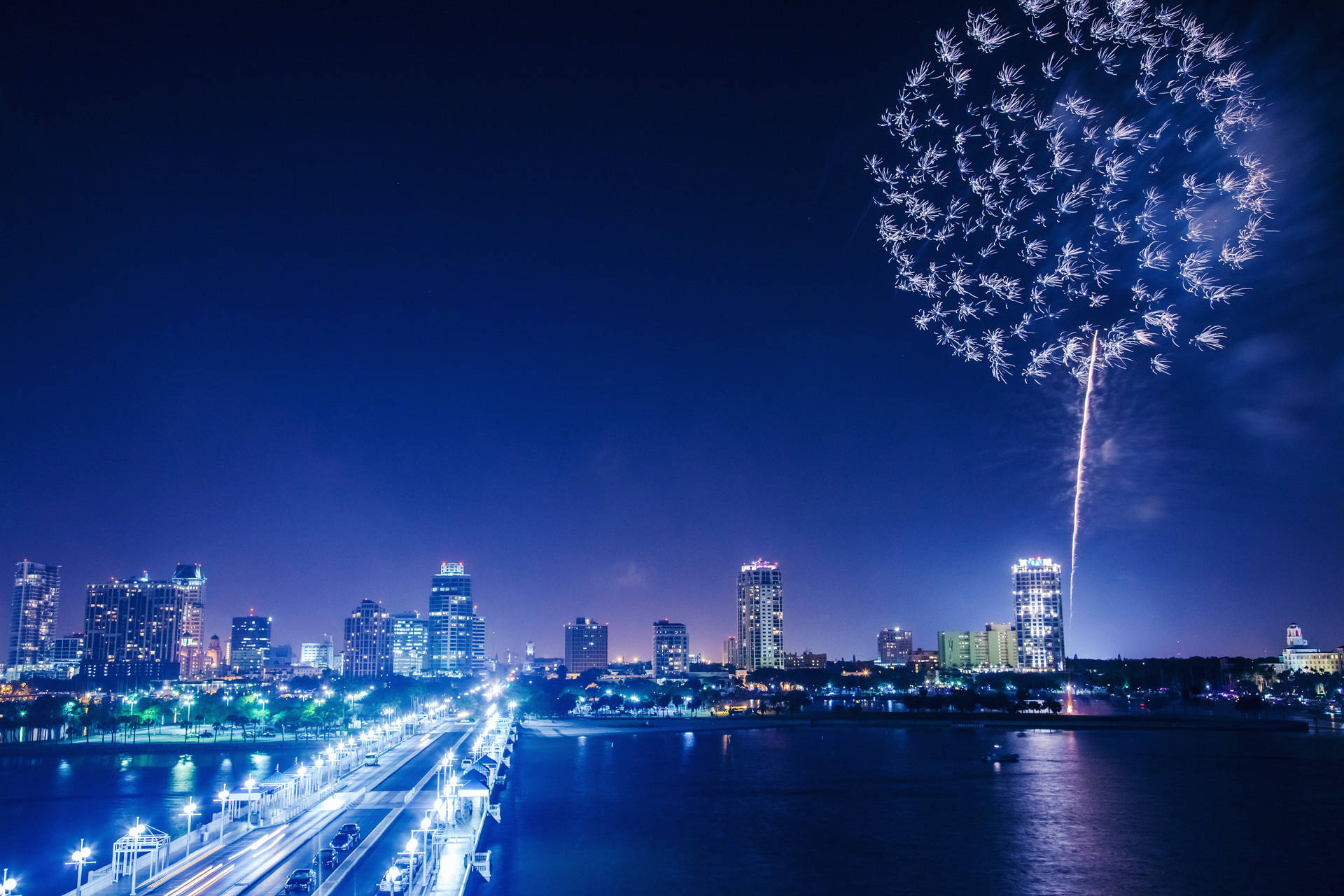 煙花在夜間城市天際線上空綻放，燈光倒映在水面上。