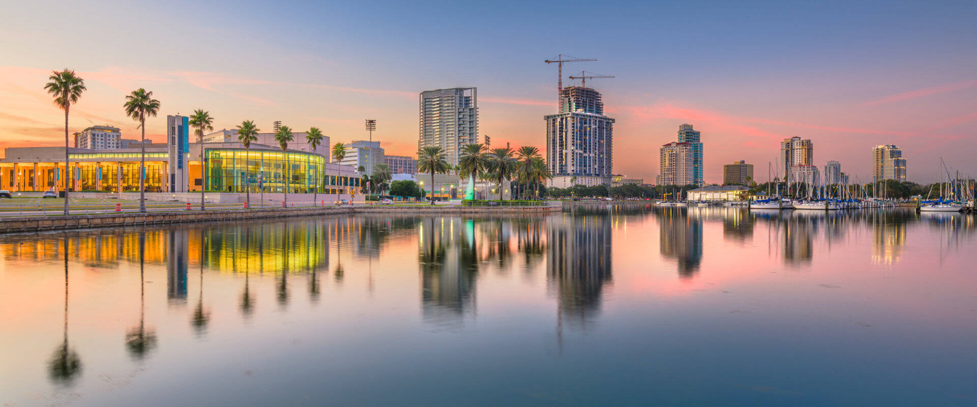 城市天際線在日落時分倒映在平靜的水面上，前景有棕櫚樹和船隻。