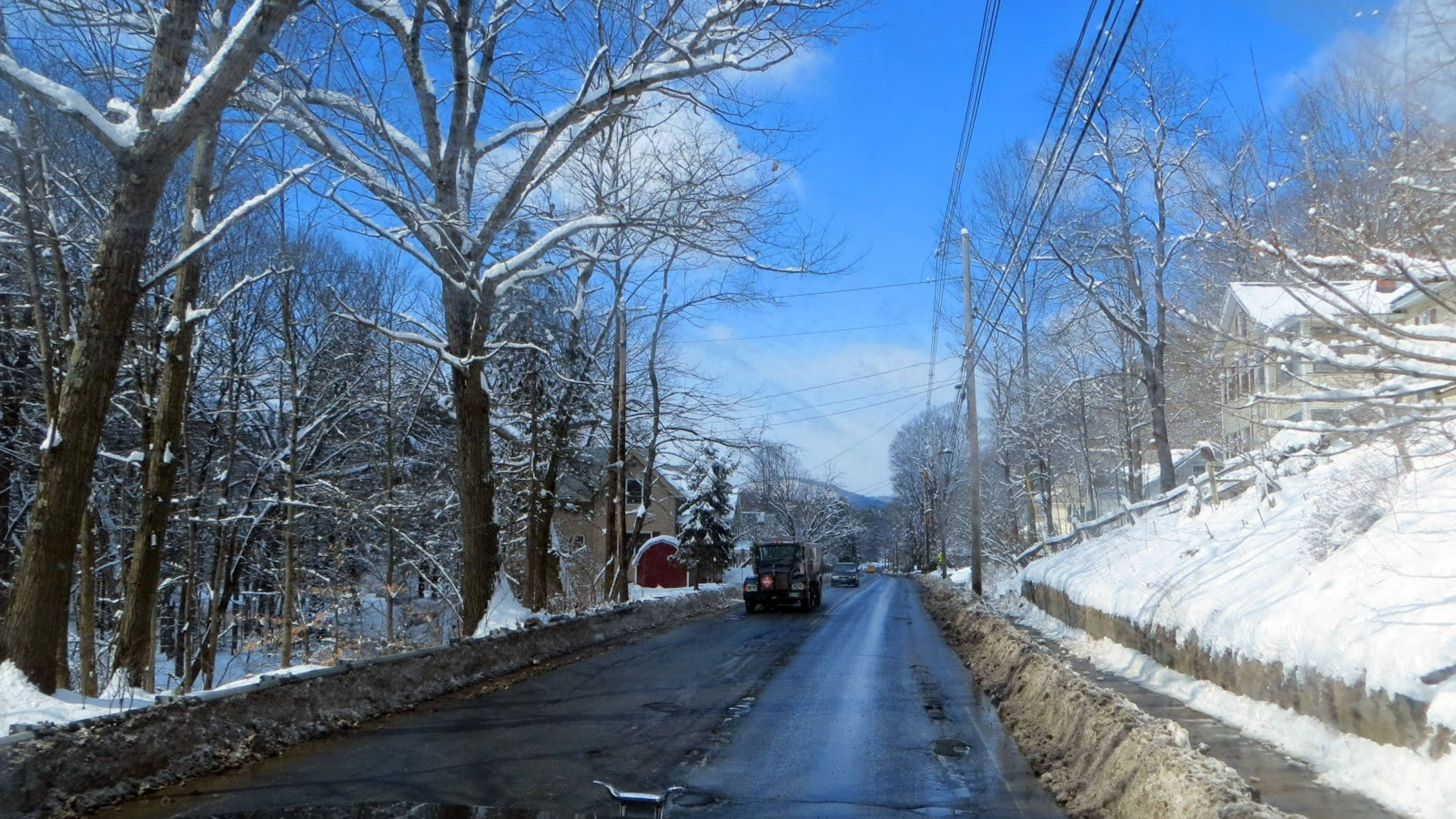 佛蒙特州冬季雪景 壁紙 — 一條積雪鄉村道路蜿蜒穿過光禿的冬季樹木，在明亮的藍天下，一輛黑色車輛駛離。