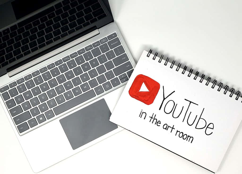 你可以成為創作者 - 在 YouTube 上傳播你的訊息！ 壁紙 — 一部銀色手提電腦和一本帶有手繪YouTube標誌的螺旋筆記本，放在潔白的表面上。