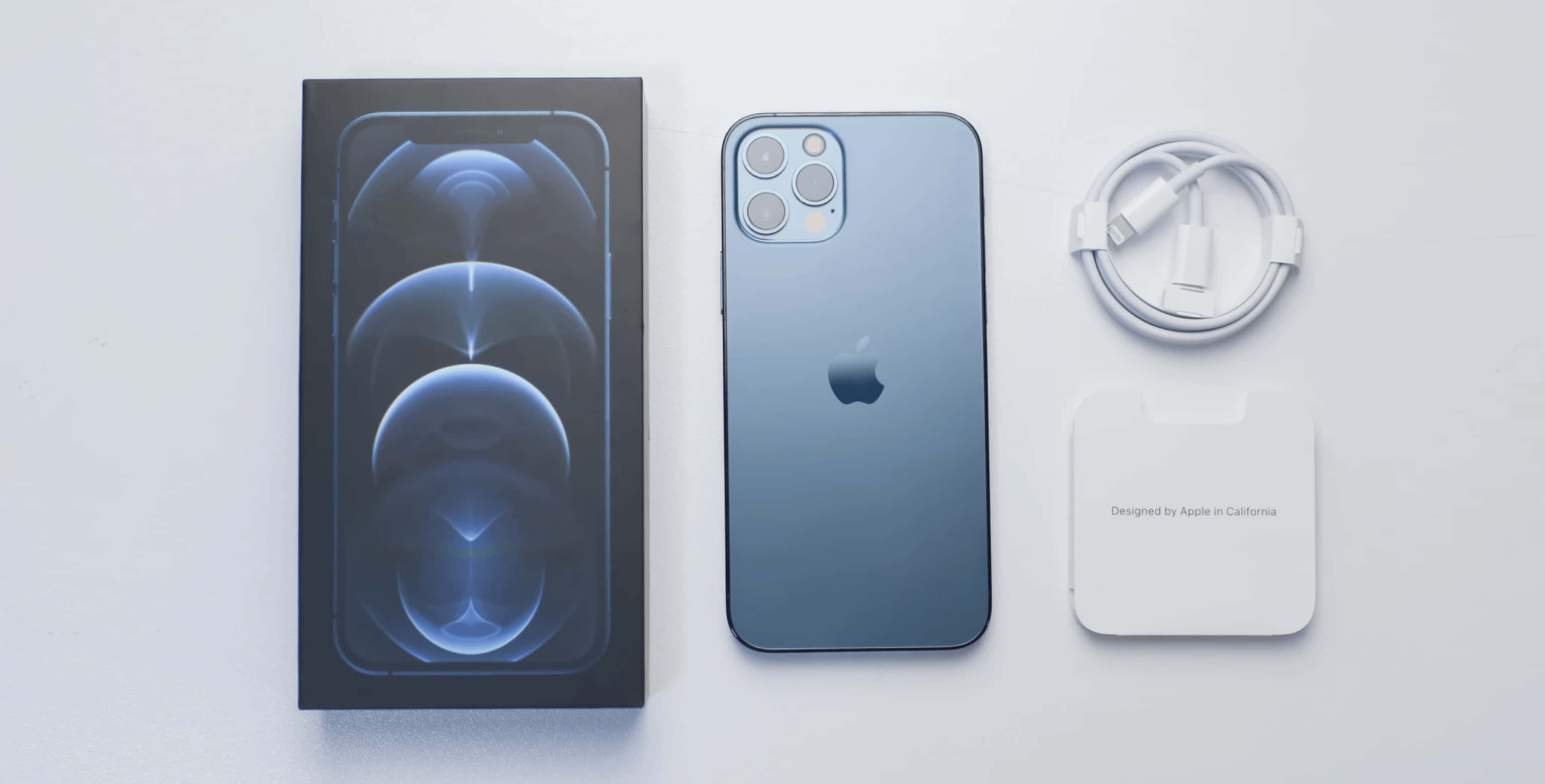 你夢寐以求的 iPhone 壁紙 — 一部時尚的藍色 iPhone 12 Pro 連同其盒裝充電器和線纜放置在簡約的白色表面上。