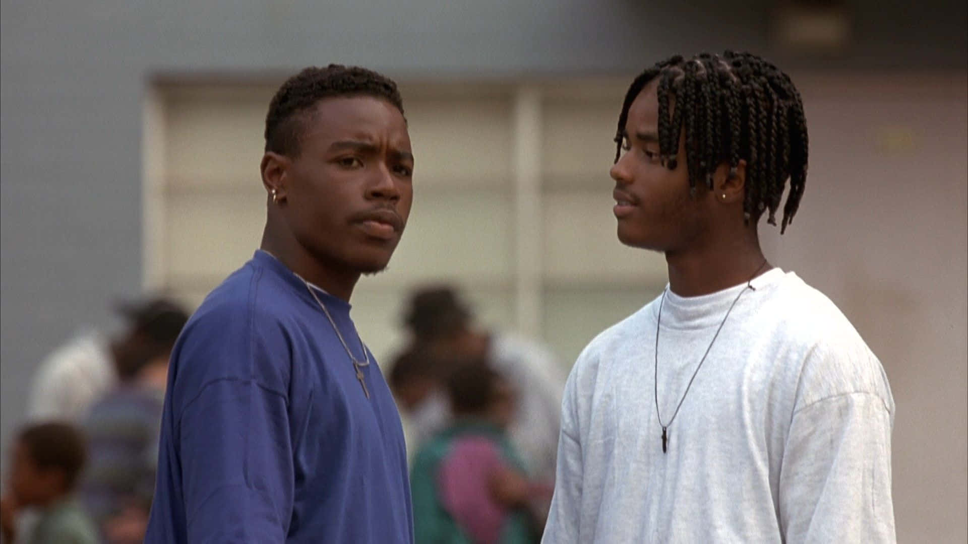 「你懂《Menace II Society》——一部關於掙扎與勝利的傳奇故事」 壁紙 — 兩位身穿休閒街頭服的年輕男子在模糊的戶外環境中面對面，表情嚴肅。