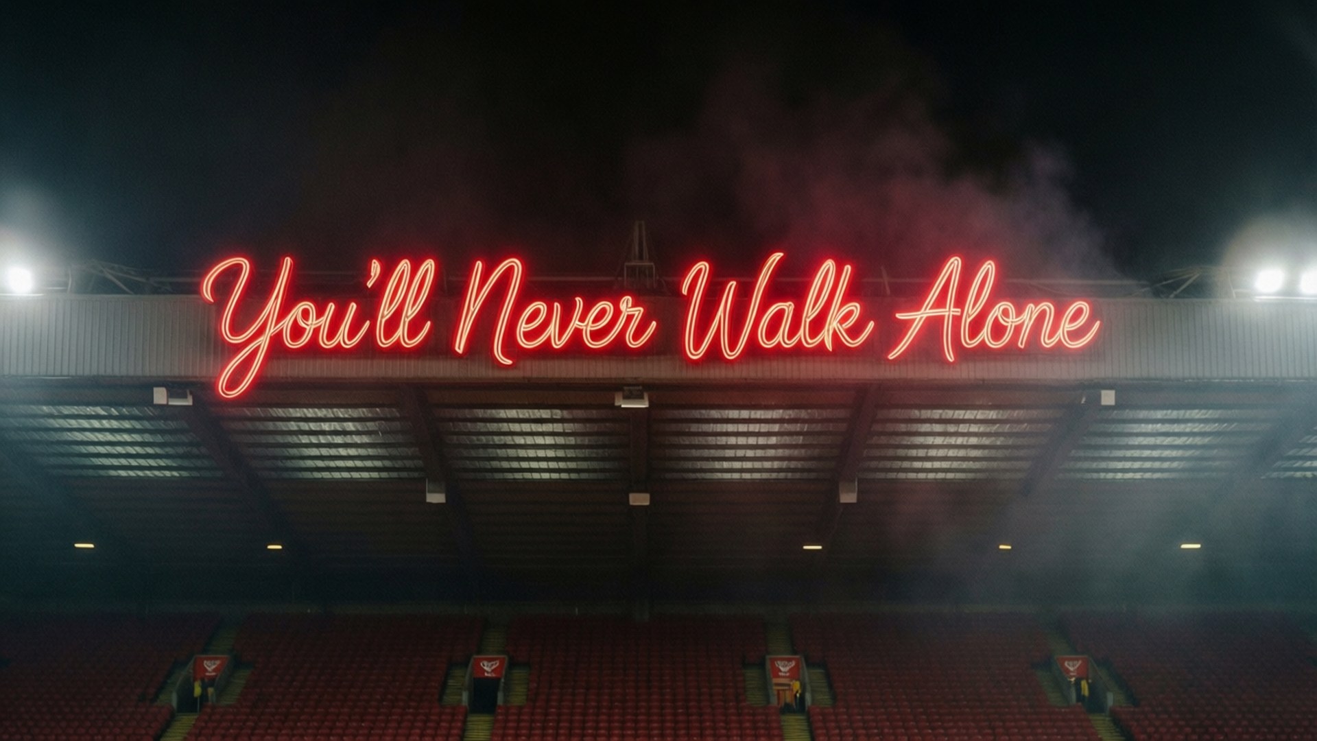 你永遠不會獨行 壁紙 — 一個發光的紅色霓虹燈牌，上面寫著 You'll Never Walk Alone，照亮了夜晚空無一人的體育場，煙霧繚繞。