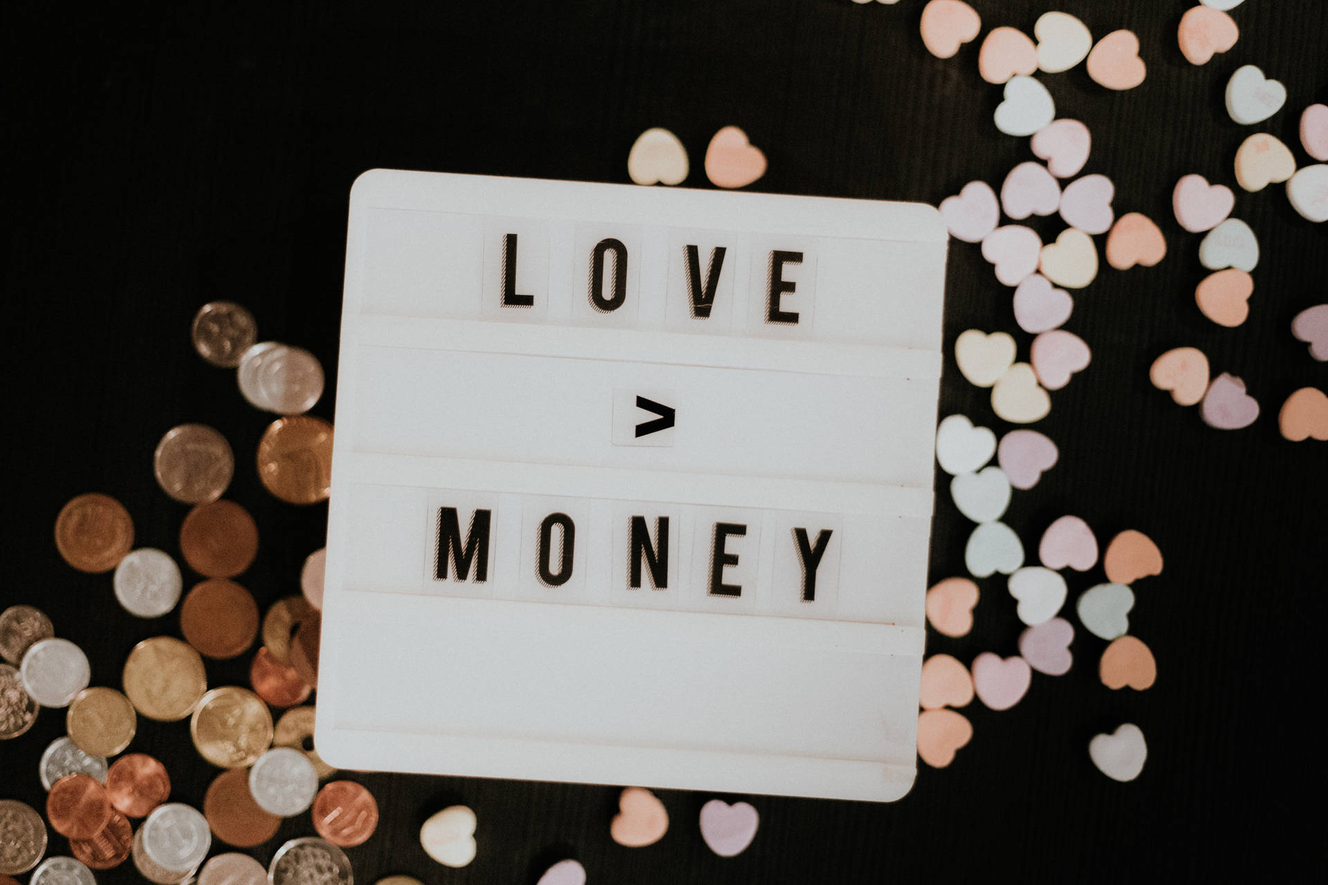白色燈箱上寫著「LOVE > MONEY」，周圍有硬幣和粉彩心形，黑色背景。